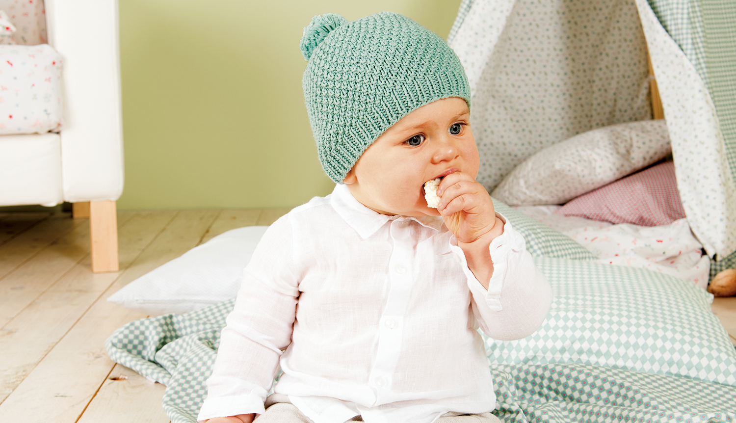 Strickanleitung Baby Mütze, Hose & Booties: Ein Baby trägt eine gestrickte Mütze und sitzt auf einer Decke.