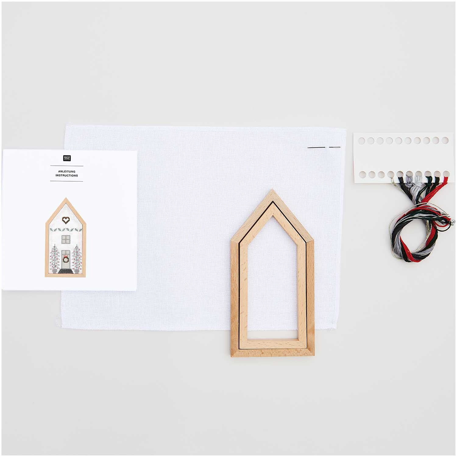 Stickpackung Haus klein 9x18cm von Rico Design mit Holzrahmen, Stoff, Anleitung und Garn auf hellem Hintergrund.