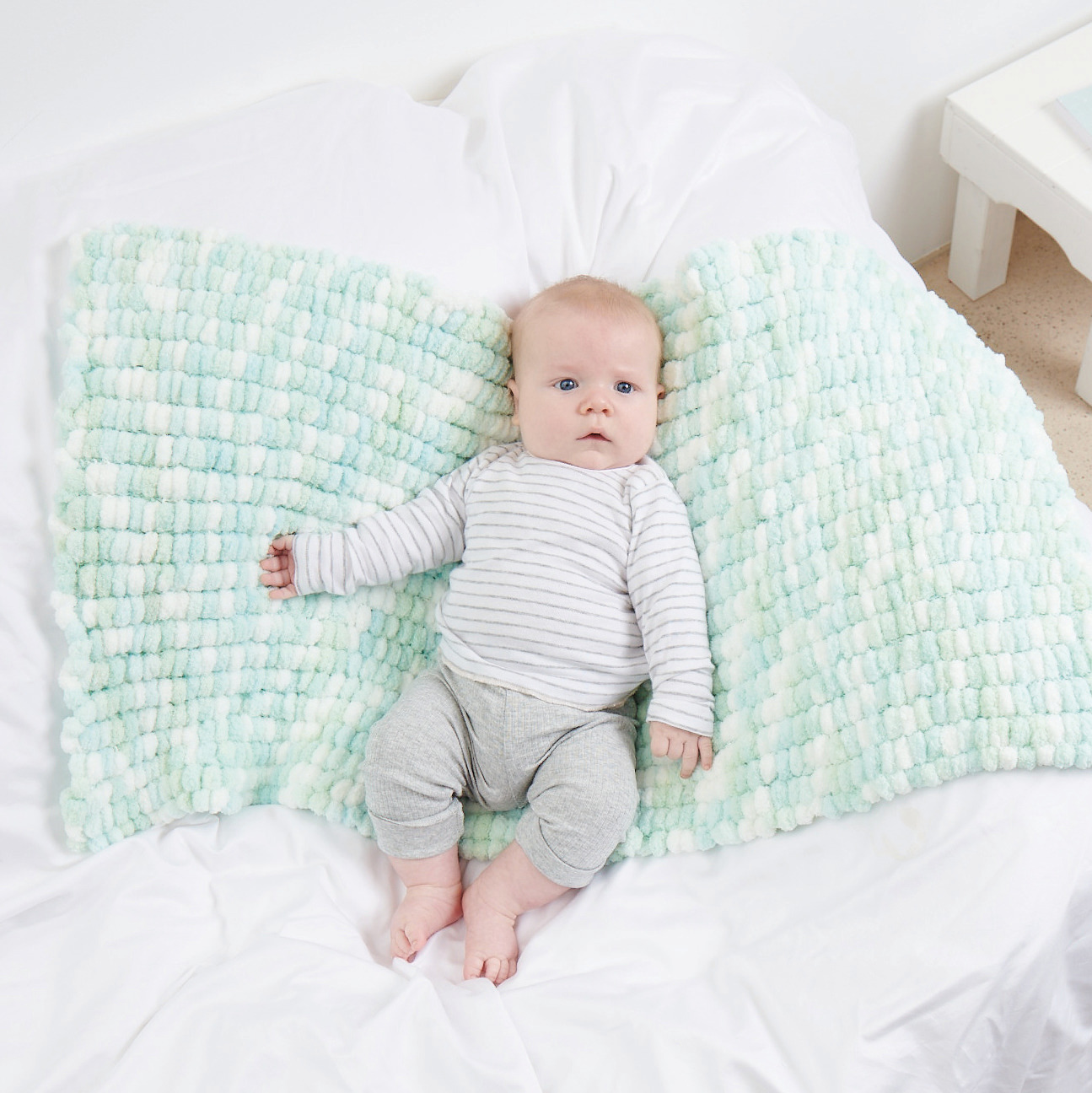 Creative Pompon Print: Baby liegt auf einer weichen, weiß-mintfarbenen Decke von Rico Design.