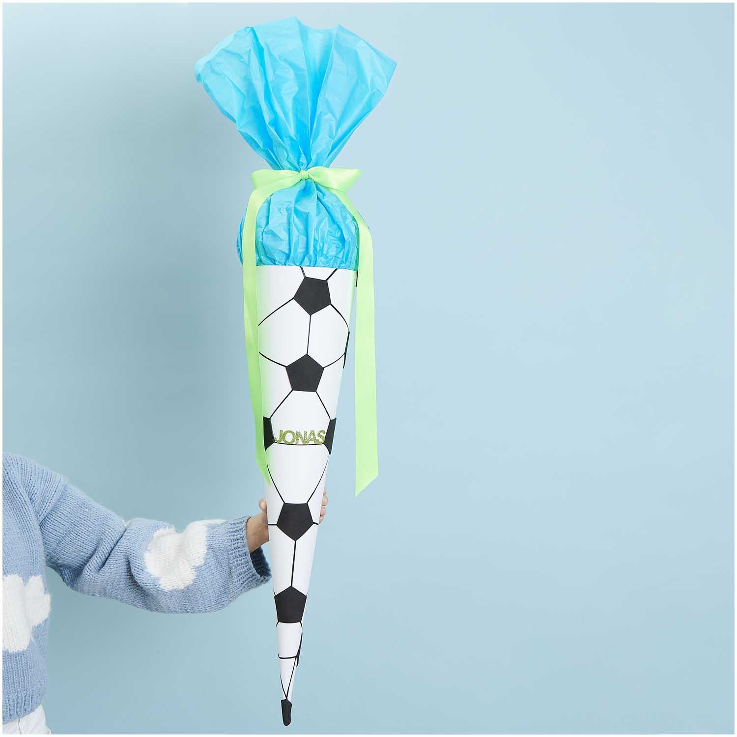 Bastelanleitung Schultüte Fussball: Kind hält eine Fußball-Schultüte mit blauem und grünem Dekor.