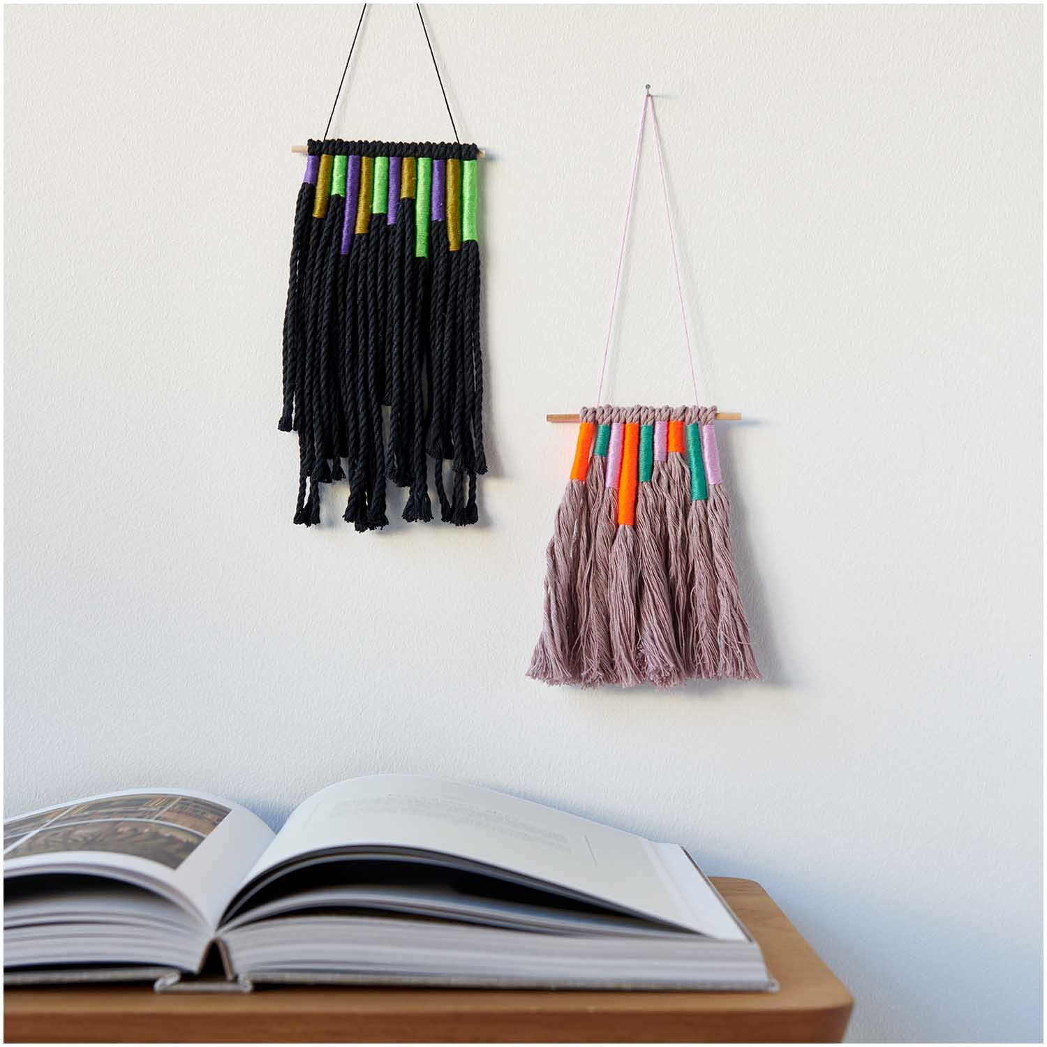 Stickgarn Set Multicolour Fashion: Zwei Wandbehänge aus buntem Garn, dekorativ über einem offenen Buch.