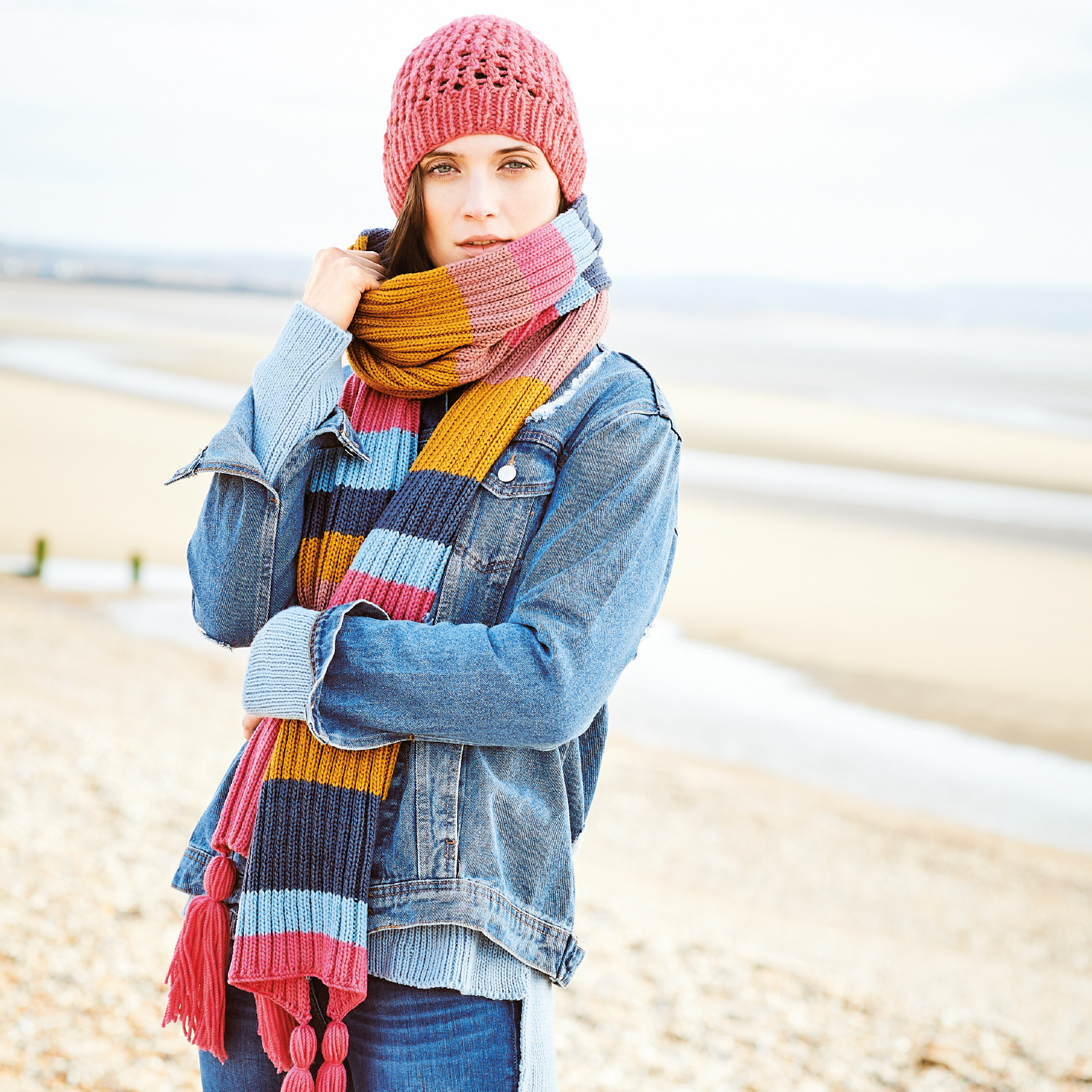 Strickanleitung Mütze und gestreifter Schal: Frau am Strand mit bunter Mütze und Schal, trägt Jeansjacke.