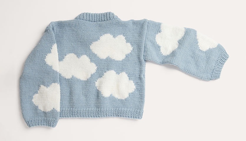 Strickanleitung Pullover aus Essentials Super aran: Hellblauer Pullover mit weißen Wolkenmotiven auf hellem Hintergrund.