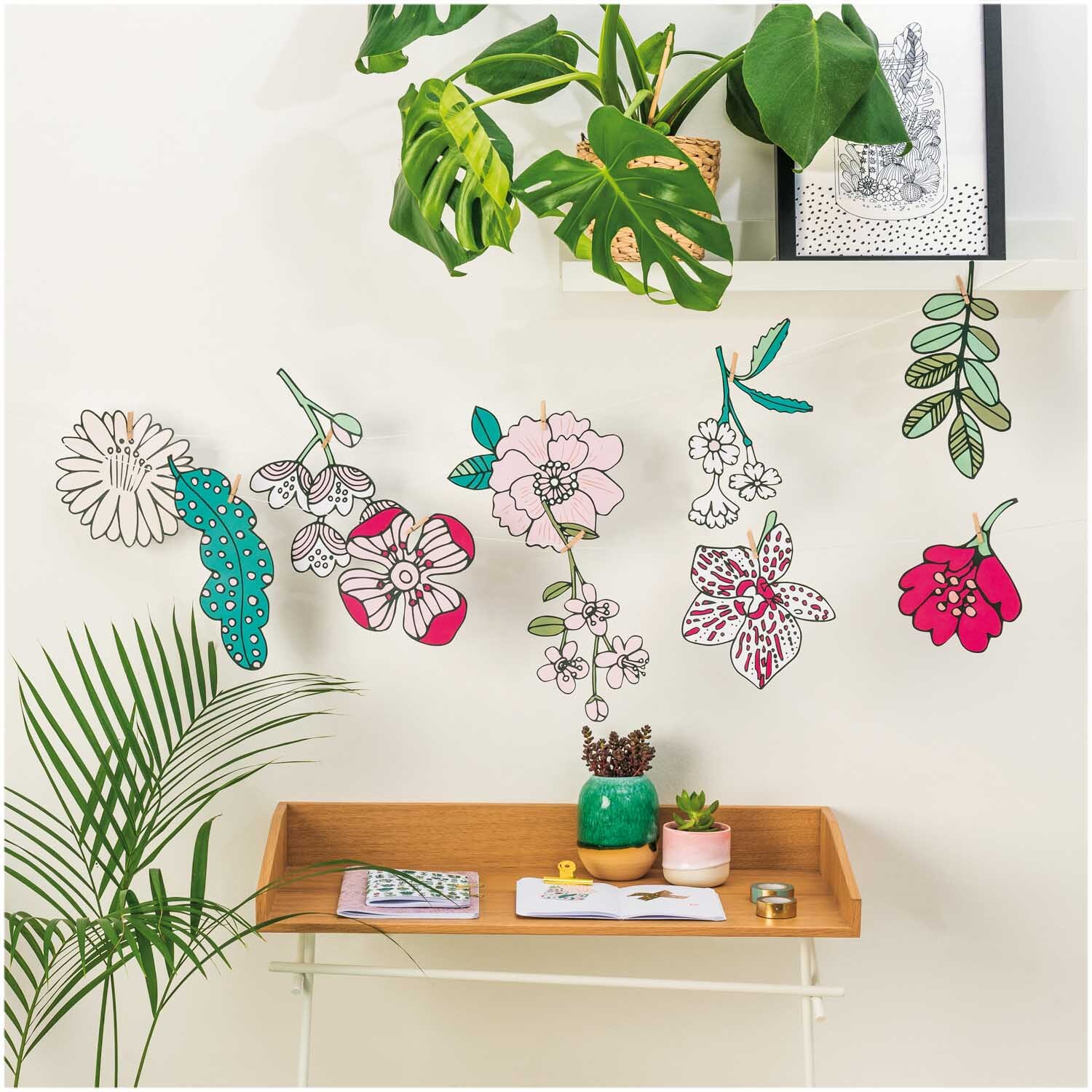 Paper Poetry Girlande Hygge Flowers 3m: Dekorative Blumen-Girlande in verschiedenen Farben von Rico Design, aufgehängt an einer Wand über einem Tisch mit Pflanzen.