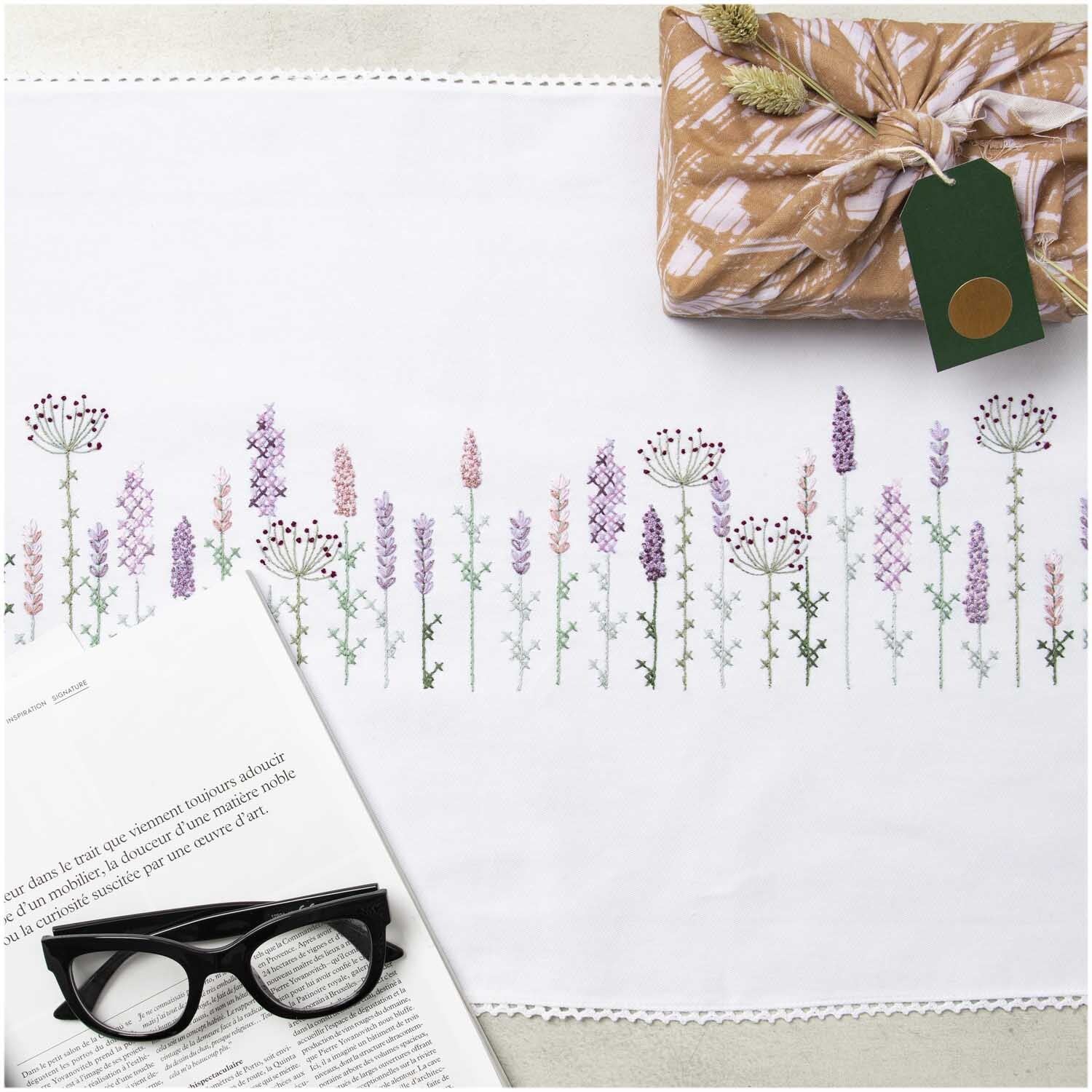 Stickpackung Läufer Lavendelfeld 45x150cm zeigt zarte Lavendelstickerei auf weißem Stoff, dekoriert mit Geschenk und Brille.