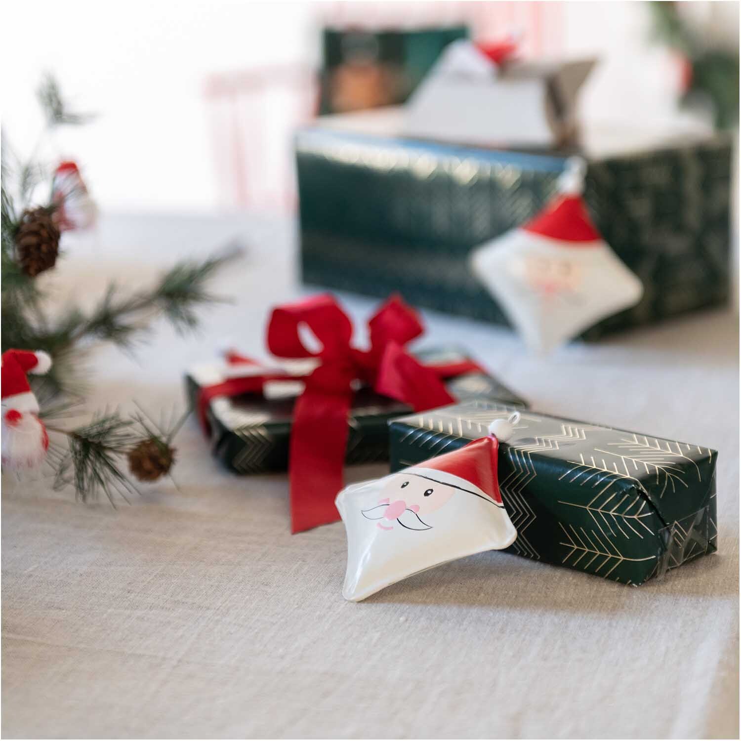 Metallhänger Weihnachtsmann rot-weiß 10x10cm, dekorativ auf Geschenken arrangiert, umgeben von Tannenzweigen.