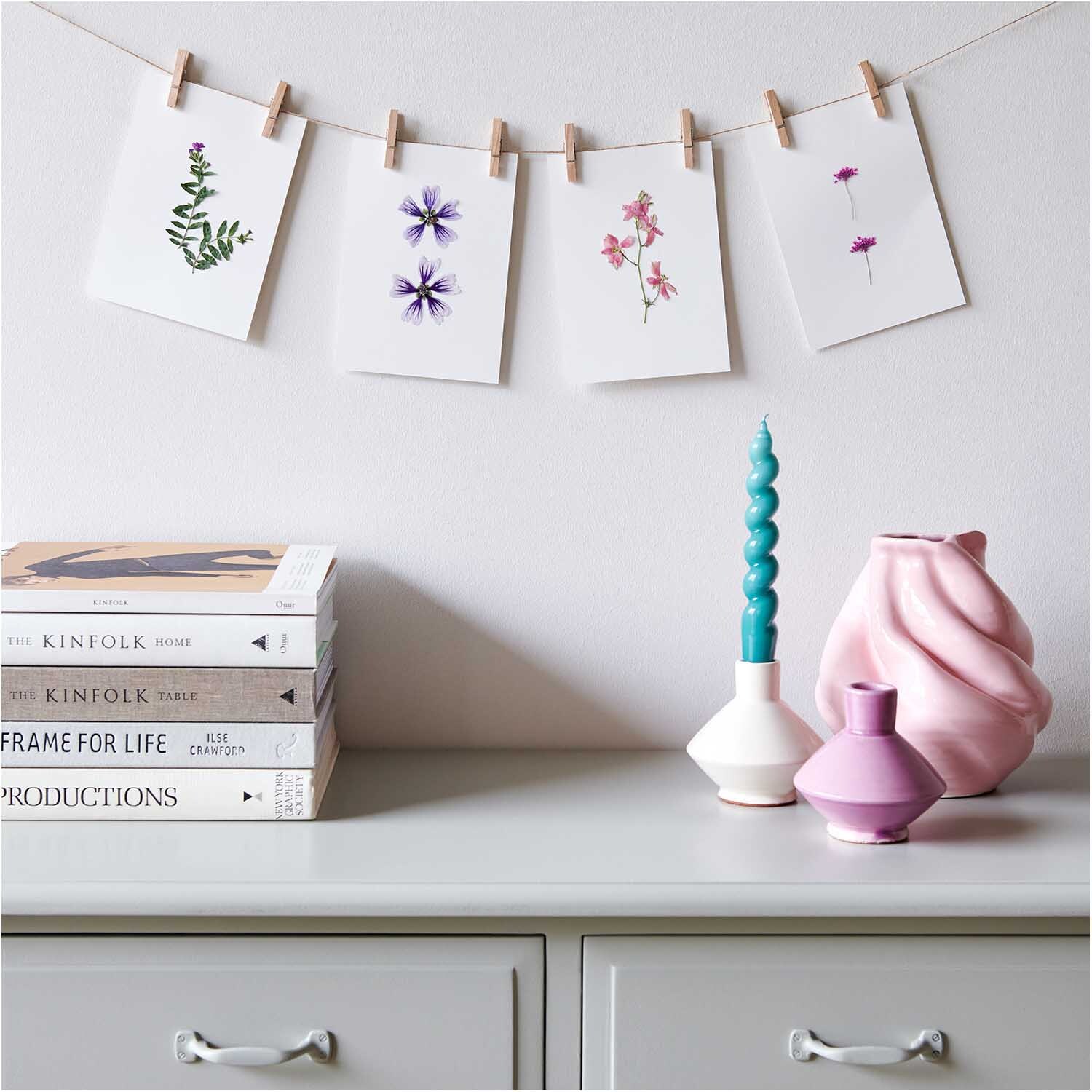 Gepresste Blüten Stockrose violett von Rico Design, dekorativ auf Karten an einer Leine präsentiert, vor einem stilvollen Regal mit Vasen und Büchern.