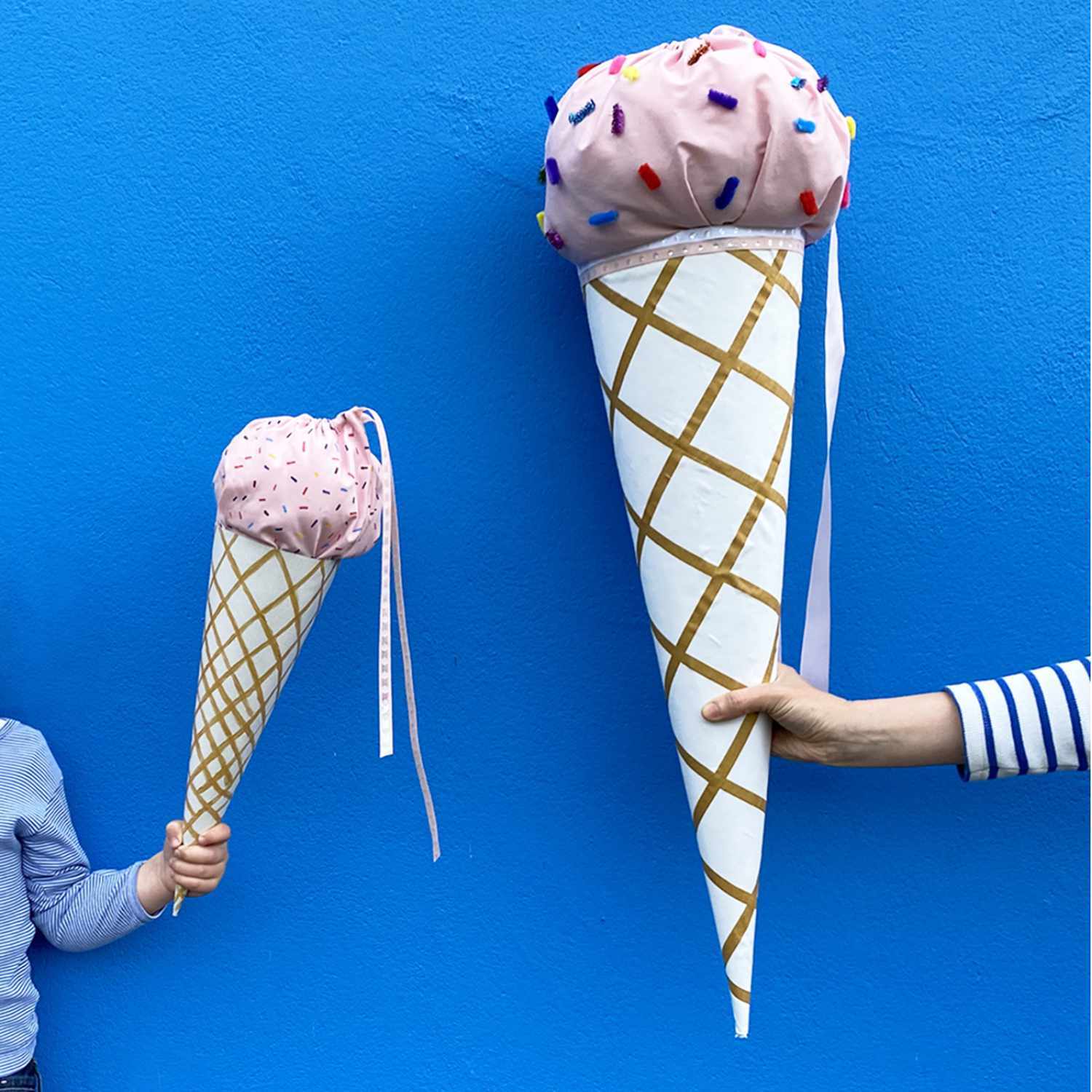 Nähanleitung Eis-Schultüten: Zwei Personen halten große, bunte Schultüten in Form von Eiswaffeln vor einer blauen Wand.