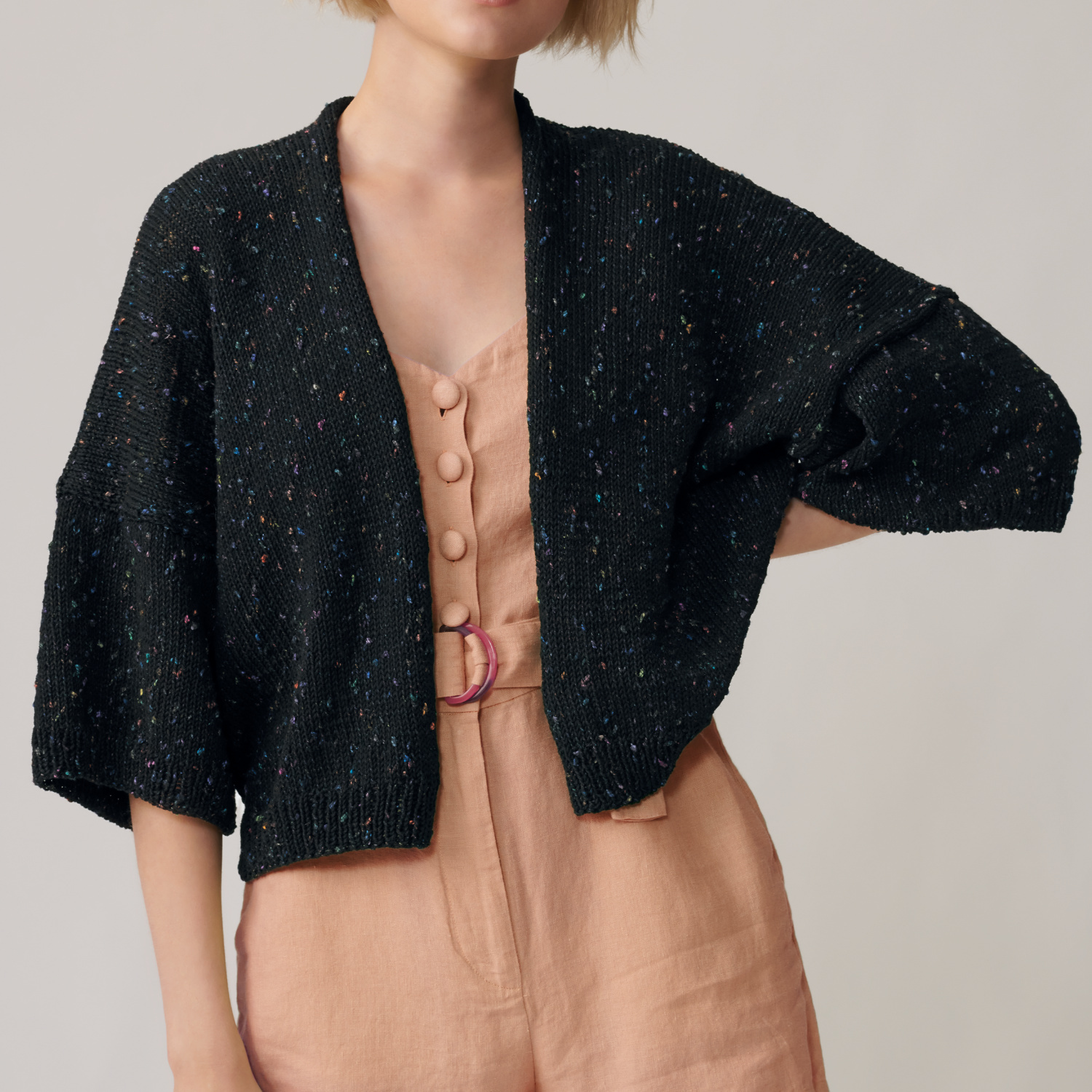 Strickanleitung Jacke aus Fashion Cotton Neps dk: Schwarze Jacke mit bunten Neps, getragen über einem beigefarbenen Jumpsuit mit Knopfleiste.