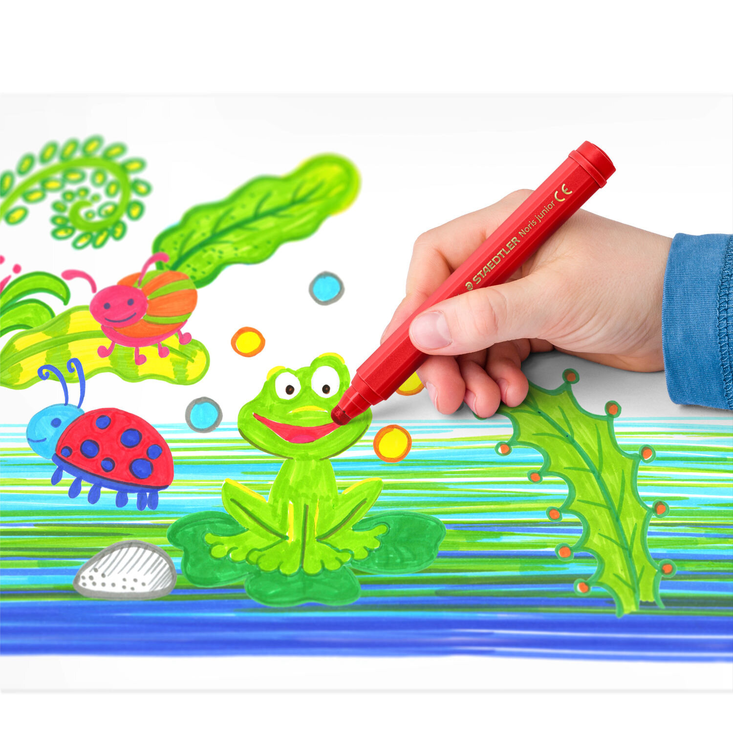 Noris Junior Fasermaler Set 6 Stück zeigt Kinderhand mit rotem Staedtler Stift, die bunten Frosch mit Käfer-Illustrationen auf weißem Papier ausmalt.