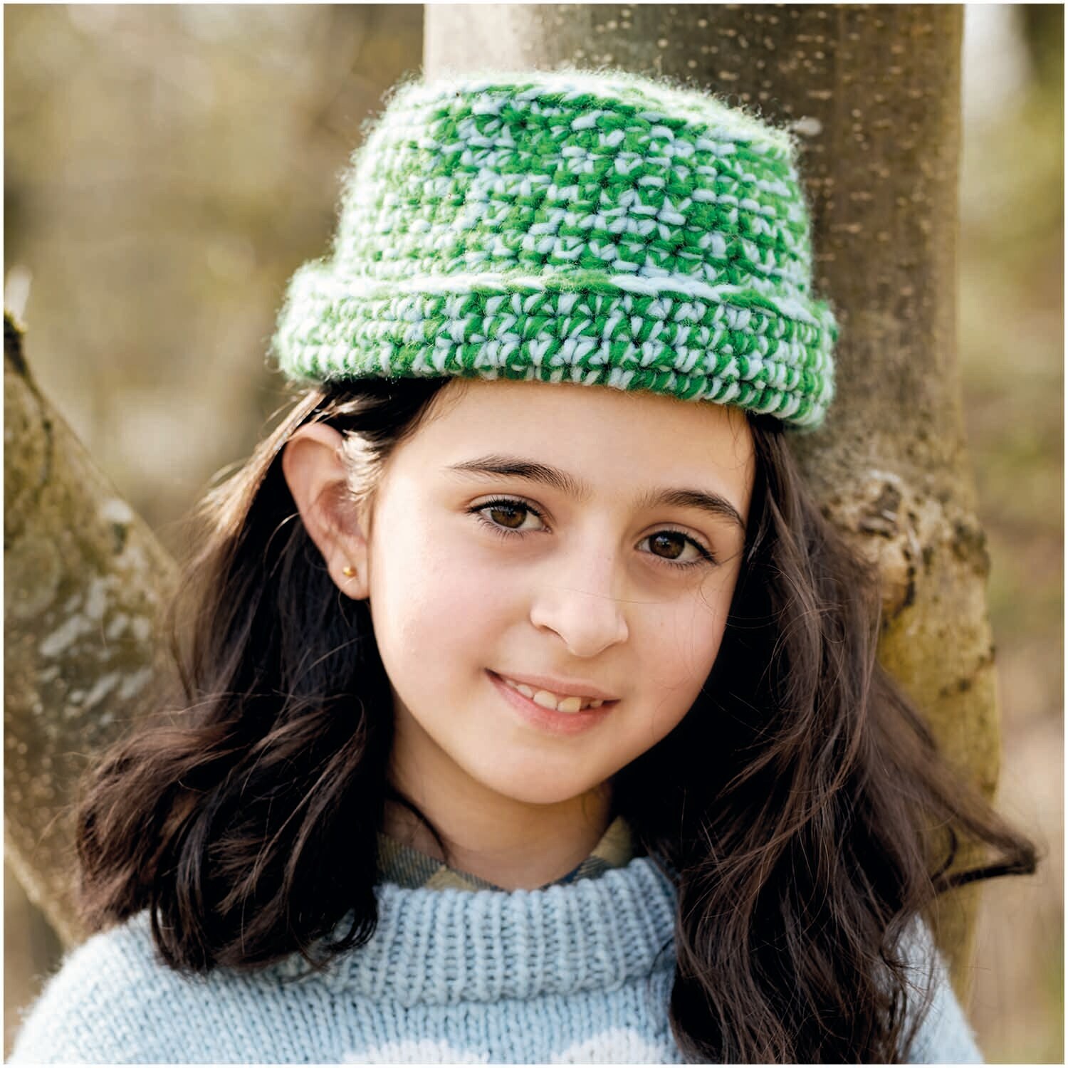 Essentials Super aran: Mädchen mit grünem, gestricktem Hut vor einem Baum, trägt hellblauen Pullover.