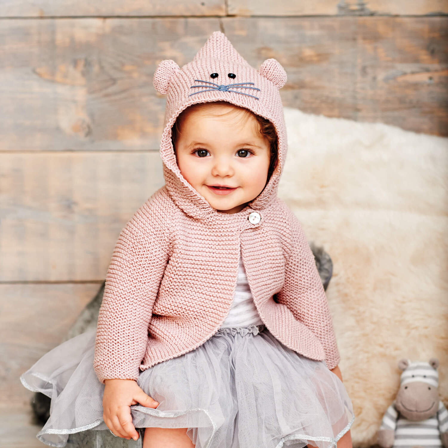 Strickanleitung Babyjacke mit Kapuze in Mausoptik: Baby trägt rosa Jacke mit Ohren, sitzt vor Holzwand.