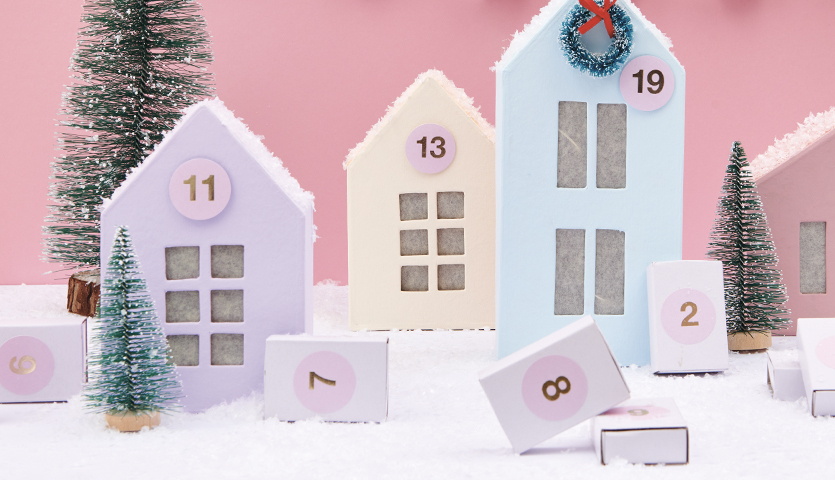 Bastelanleitung Adventskalender Pappmaché-Häuser: Pastellfarbene Häuser mit Zahlen auf Schnee.