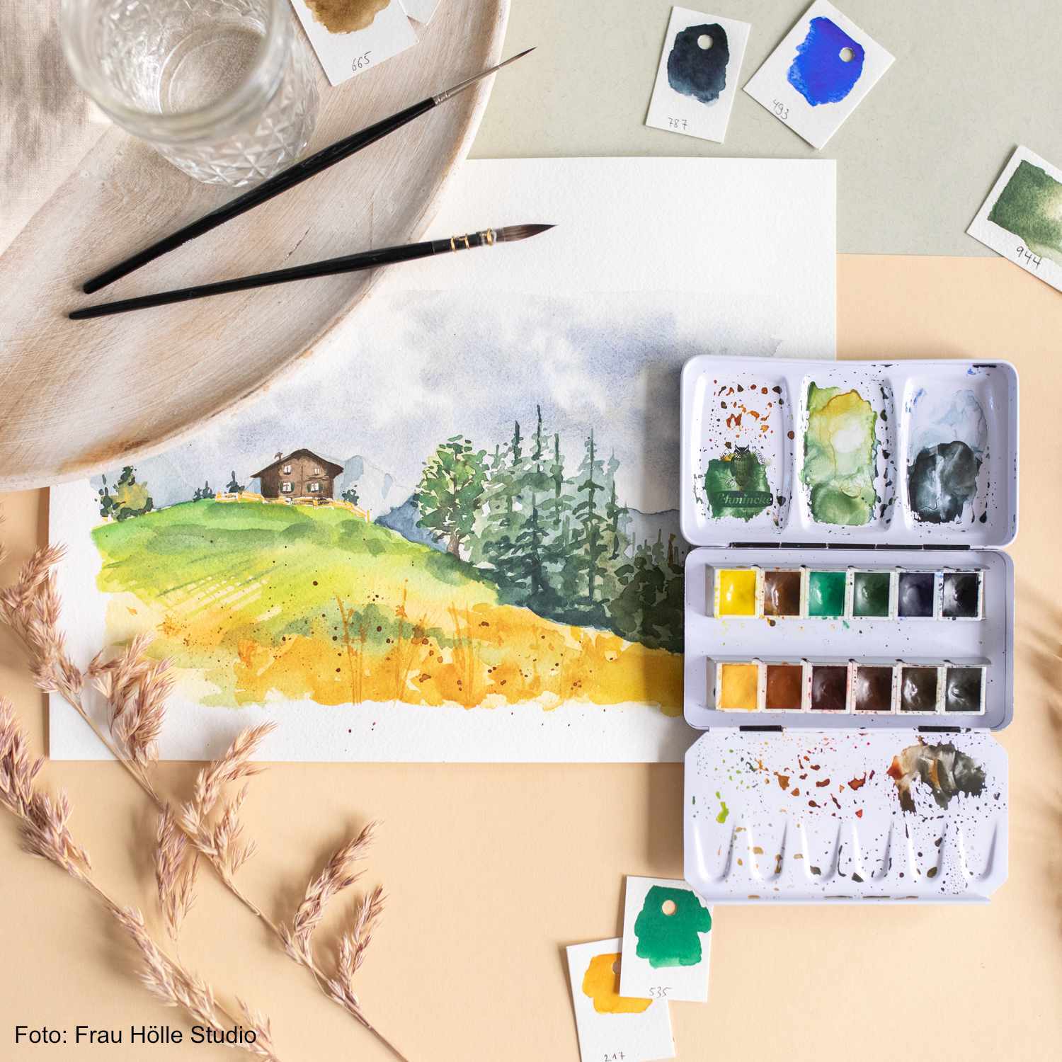 Schmincke Horadam Aquarellkasten mit 12 Farben, ideal für Landschaftsmalerei, neben einem Aquarellbild einer Hügellandschaft.