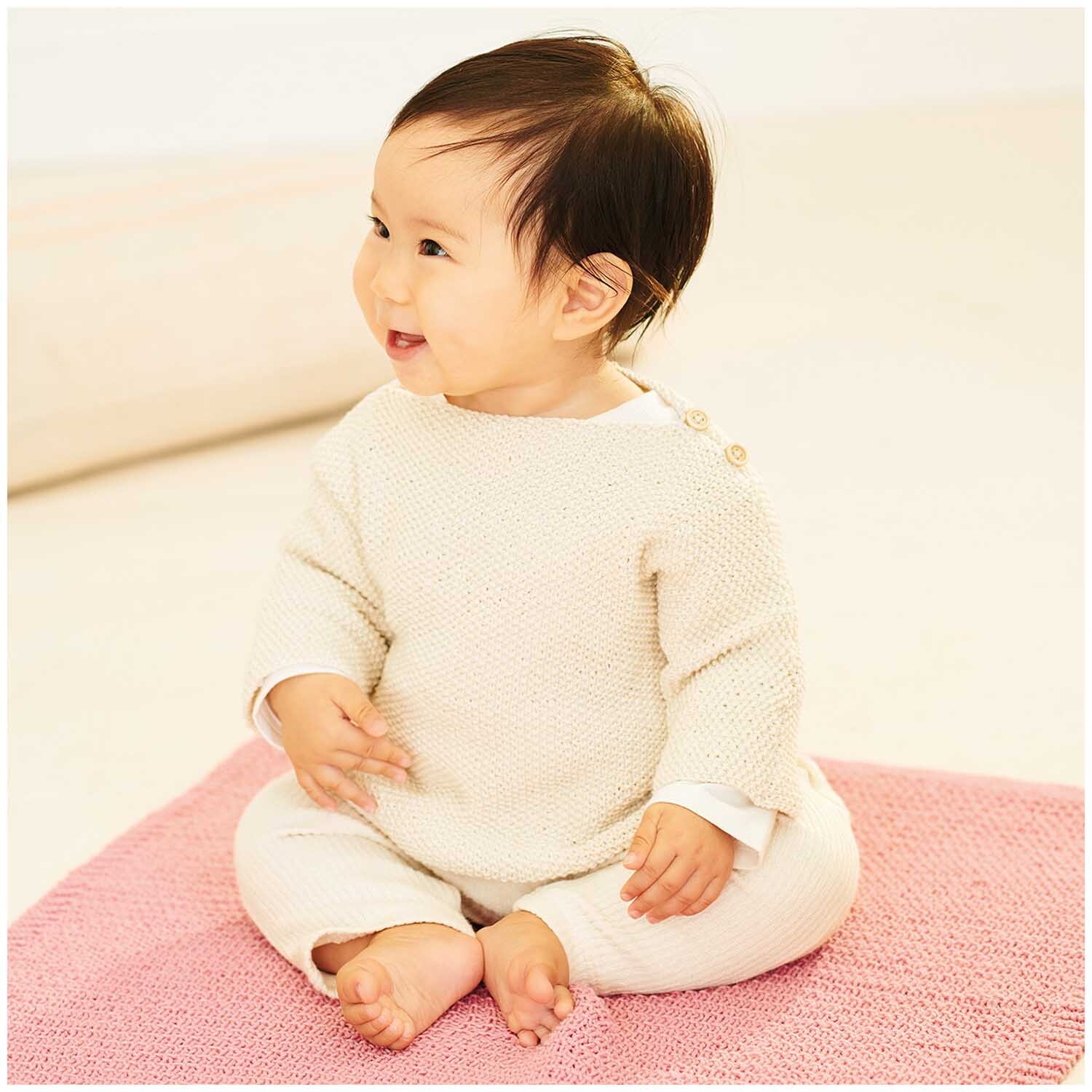 Baby Organic Cotton: Ein lächelndes Baby in cremefarbener Kleidung sitzt auf einer rosa Decke.