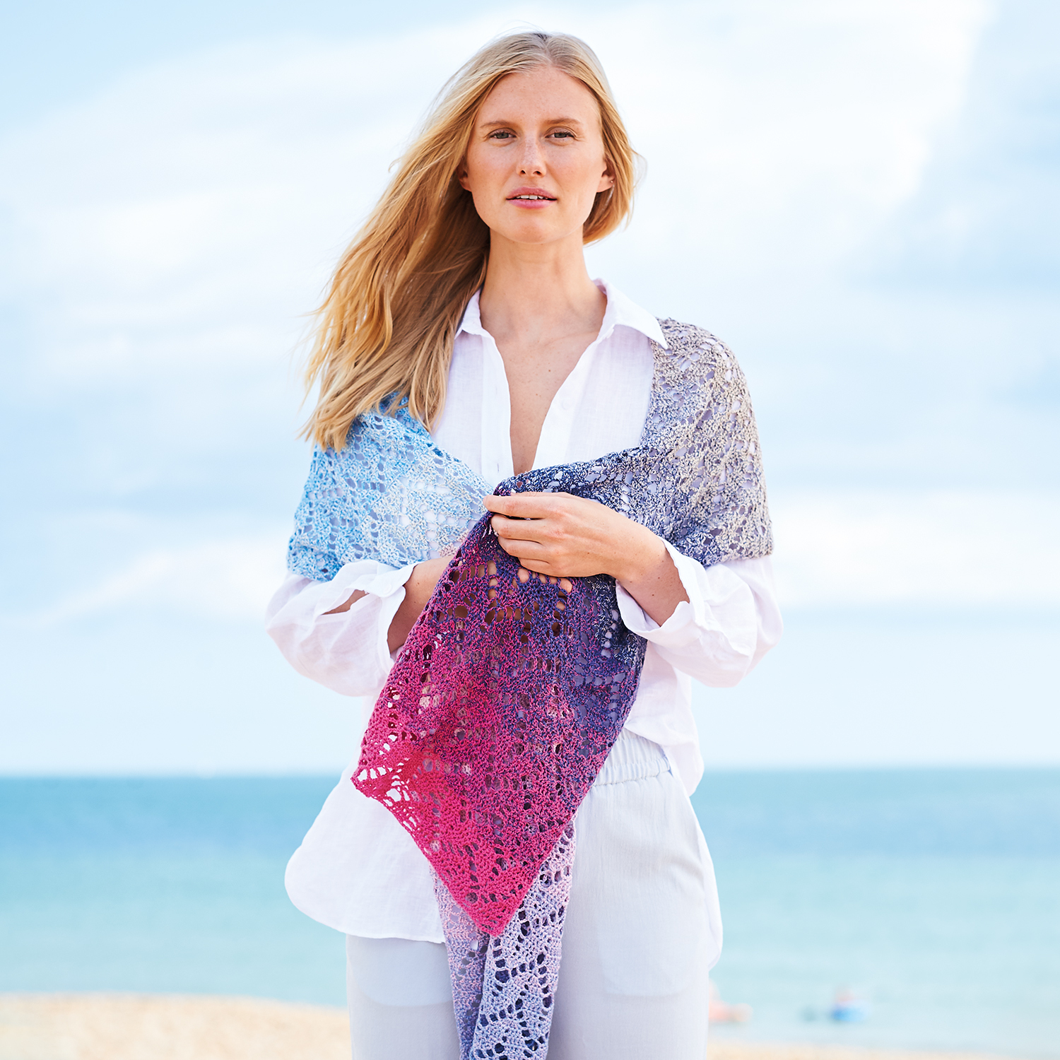 Häkelanleitung Tuch aus Creative Cotton Dégradé Lucky 8: Frau am Strand mit farbenfrohem, gehäkeltem Tuch in Blau- und Rottönen.
