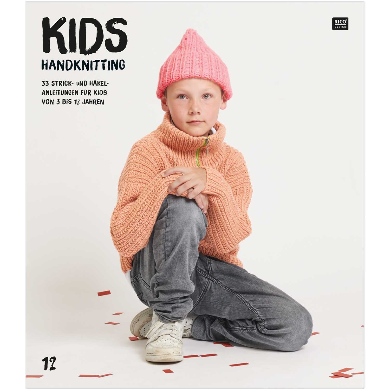 Kids Nr.12: Kind in rosa Mütze und orangefarbenem Pullover sitzt auf dem Boden, umgeben von roten Konfetti.