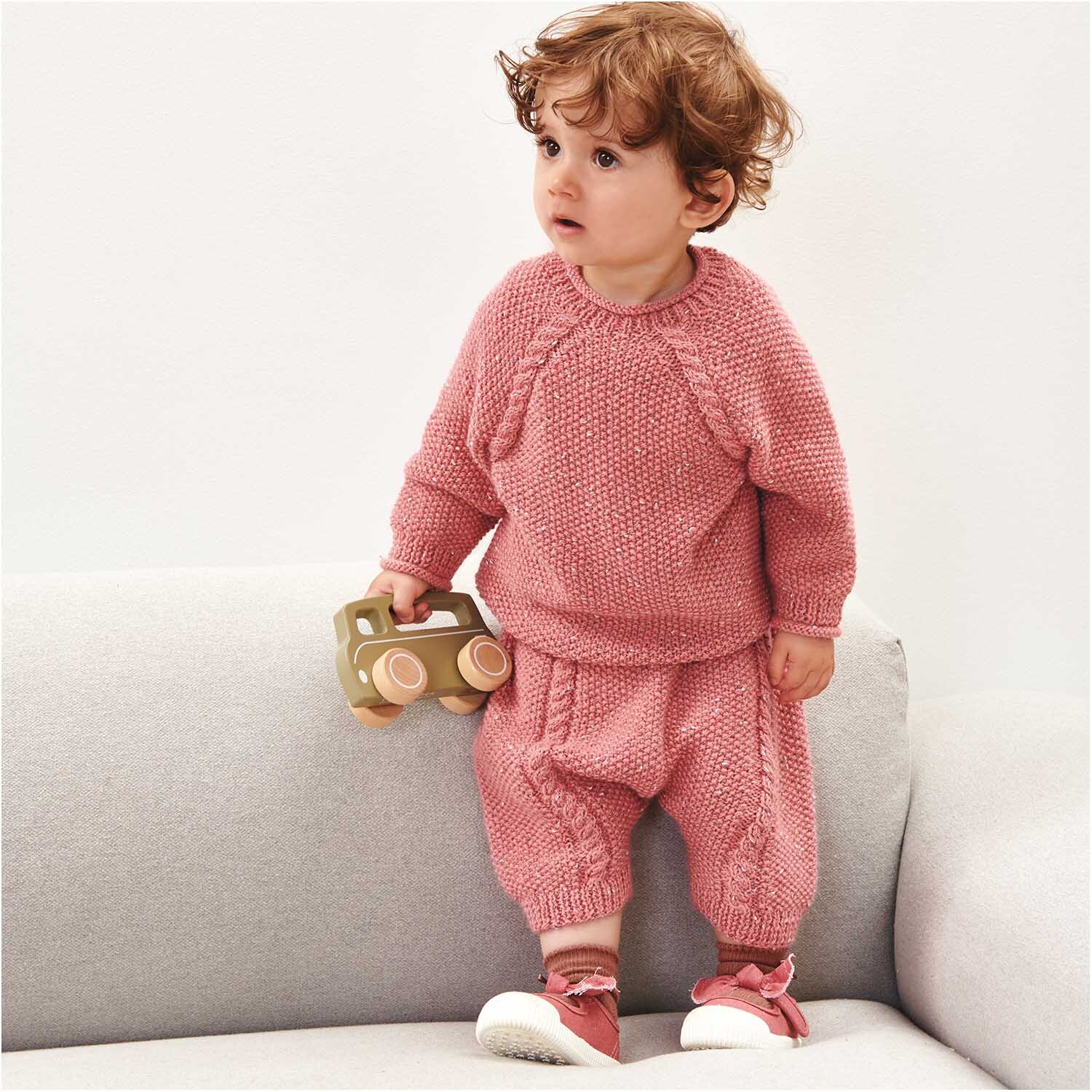 Rico Baby Dream Tweed dk - A Luxury Touch: Kind in gestricktem Set in Azalee auf Sofa mit Spielzeug.