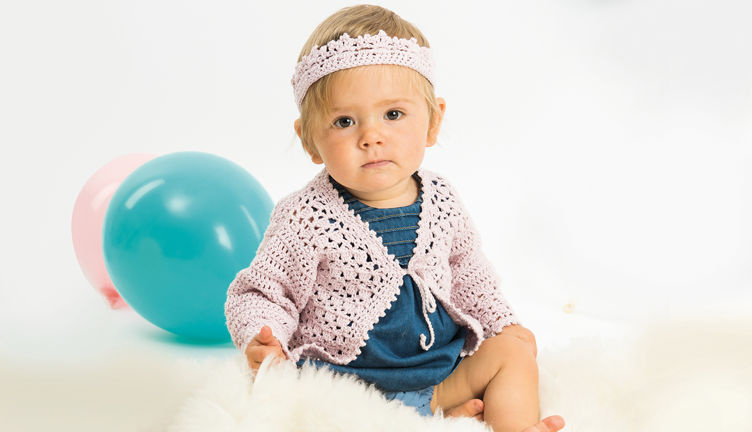 Häkelanleitung Baby-Jacke mit Krone: Ein Baby trägt eine gehäkelte Jacke und Krone, sitzt auf einem weißen Teppich mit bunten Luftballons im Hintergrund.