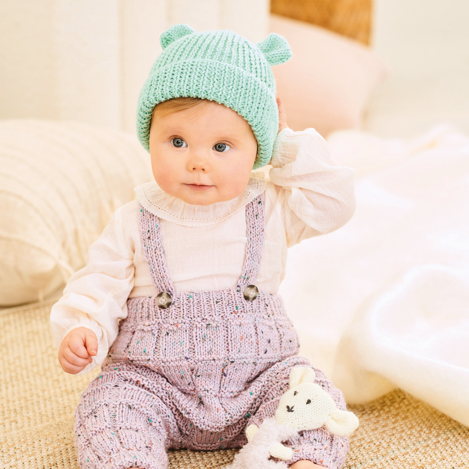 Strickset Hose Modell 07 aus Rico Baby 040: Baby in lila Strickhose und mintgrüner Mütze auf beigem Teppich.