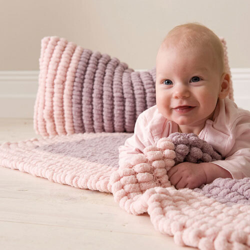 Pompon: Ein Baby liegt auf einer weichen, pastellfarbenen Decke von Rico Design, umgeben von Kissen.