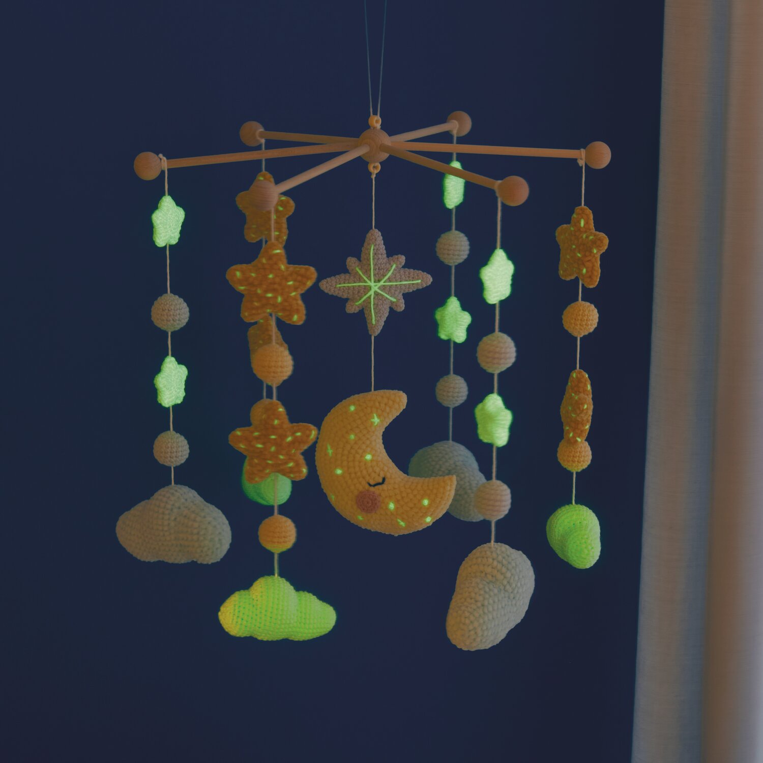 Ricorumi Glow In The Dark Mobile mit leuchtenden Sternen und Mond, gehäkelt in Eisgrün und Silbergrau, hängt vor dunkelblauem Hintergrund.
