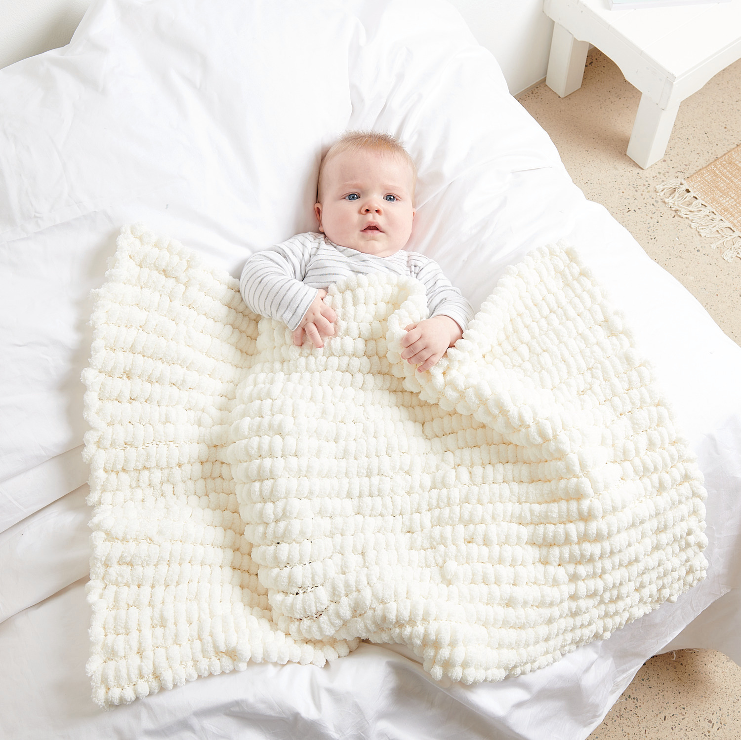 Pompon: Baby in cremefarbener Decke aus weicher Wolle, gemütlich auf weißem Bett liegend.