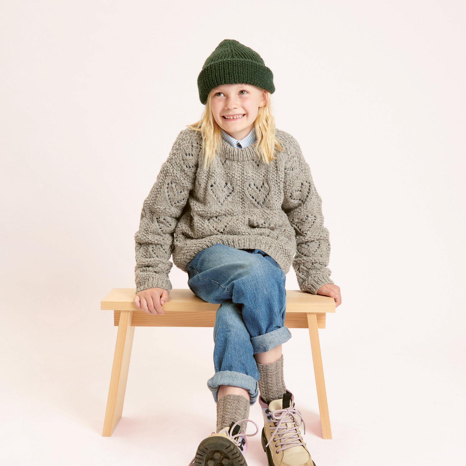 Strickset Kinderpullover Modell 06: Kind in grauem Pullover und olivgrüner Mütze auf Holzbank.