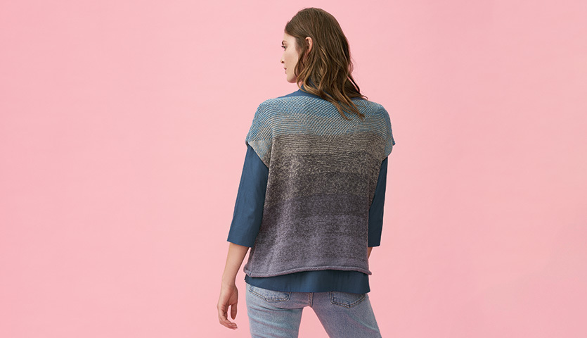 Strickanleitung Top aus Creative Cotton Dégradé Super 6: Frau trägt blau-graues Stricktop vor rosa Hintergrund, kombiniert mit Jeans.