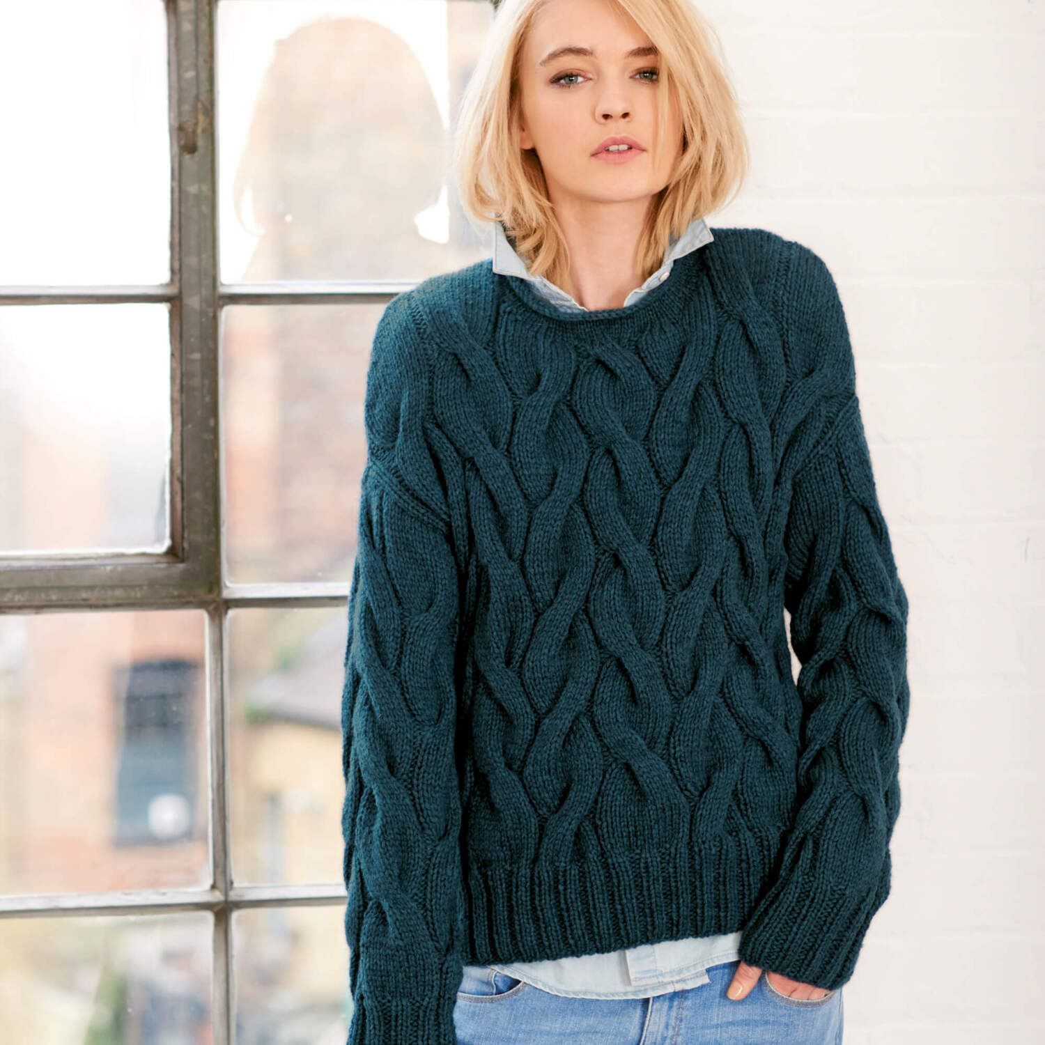 Strickanleitung Pullover mit groben Zöpfen: Frau trägt blauen Zopfpullover vor einem Fenster.