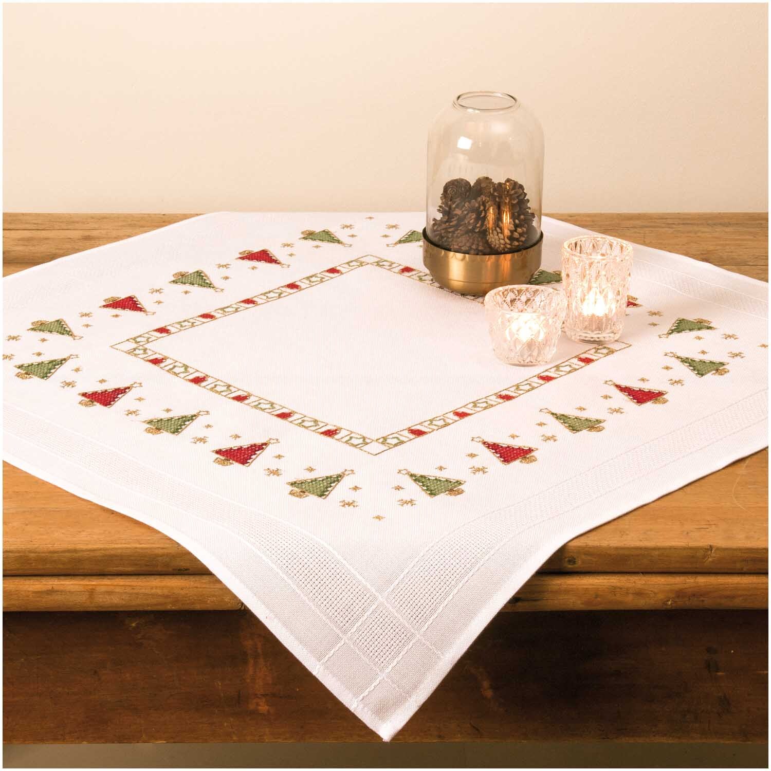 Stickpackung Tischdecke Tannenbordüre 80x80cm auf Holztisch, dekoriert mit Glasglocke und Kerzen, von Rico Design.
