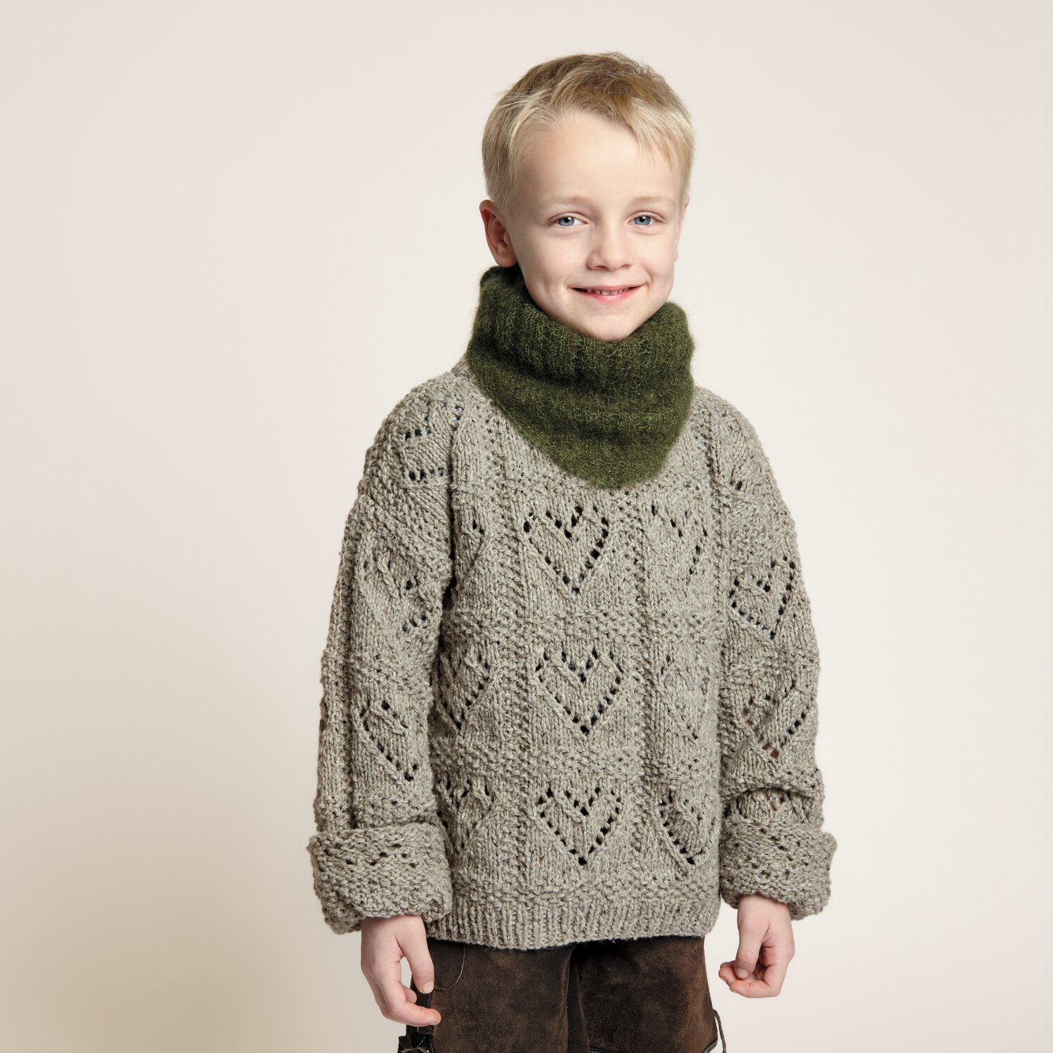 Strickset Kinderpullover Modell 06: Junge trägt gestrickten Pullover mit Herzmuster und olivgrünem Schal. Ideal für Trachtenmode von Rico Design.