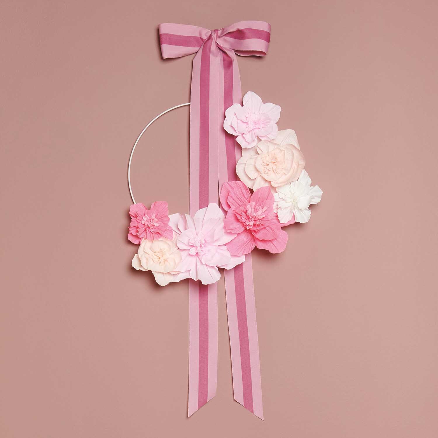 Bastelanleitung Blumenkranz aus Krepppapierblumen: Ein Kranz mit rosa und weißen Papierblumen und einer gestreiften Schleife auf rosa Hintergrund.