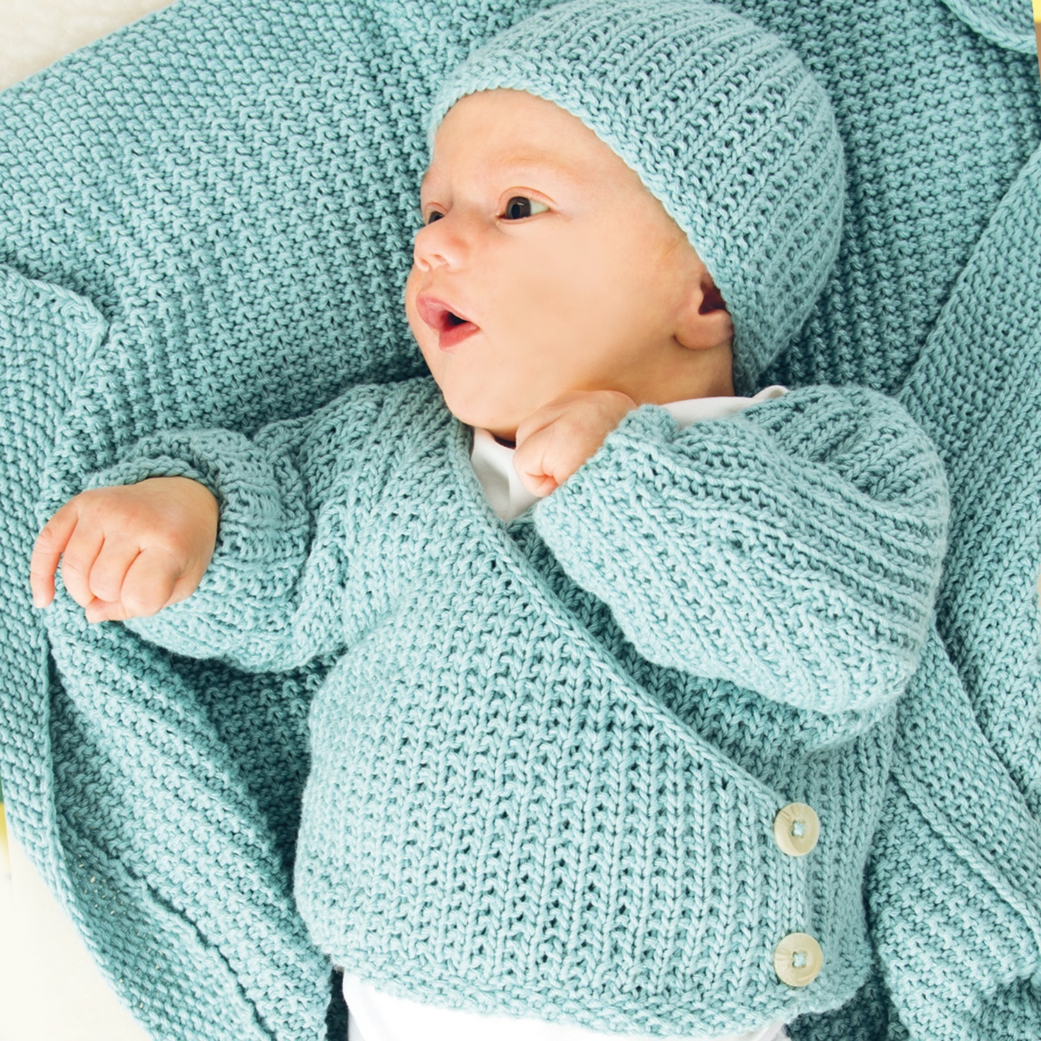 Strickanleitung Baby-Jacke mit Knöpfen: Baby in blauer Strickjacke und Mütze auf passender Decke.
