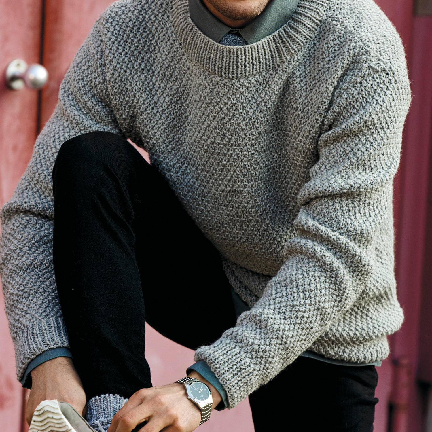 Strickanleitung Herrenpullover im Perlmuster: Mann trägt grauen Pullover aus Essentials Soft Merino aran, sitzt auf einer Treppe.