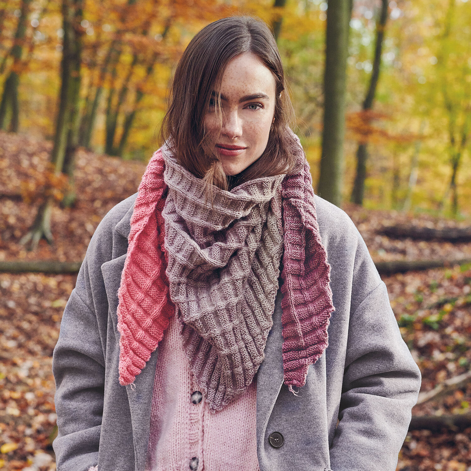 Strickanleitung Dreieckstuch Creative Wool Dégradé Super 6: Frau im Wald mit farbigem, gestricktem Dreieckstuch, das in Rosa- und Brauntönen gehalten ist.