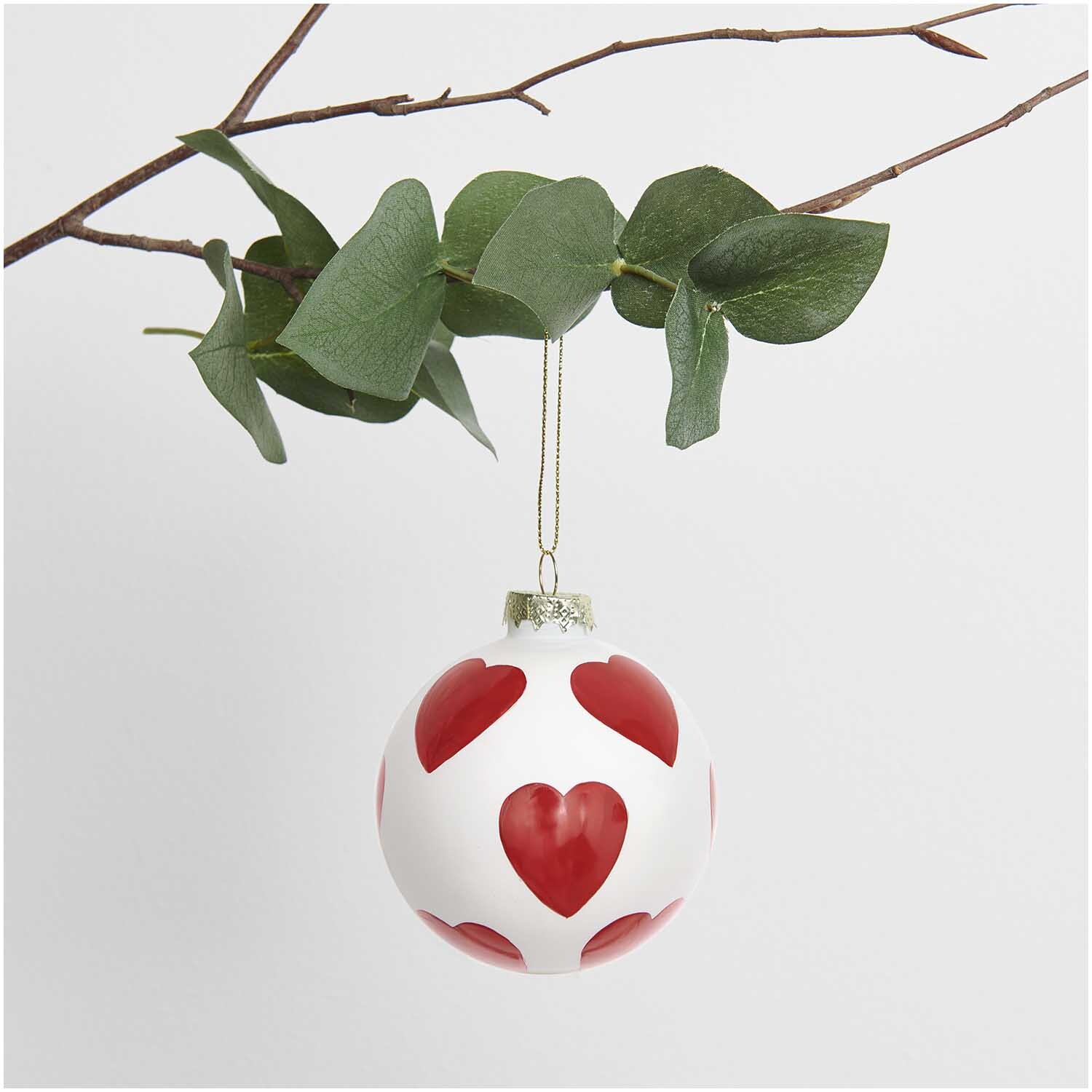 Glaskugel Baumschmuck in Herzform, hängend an einem Ast mit grünen Blättern, von Rico Design.