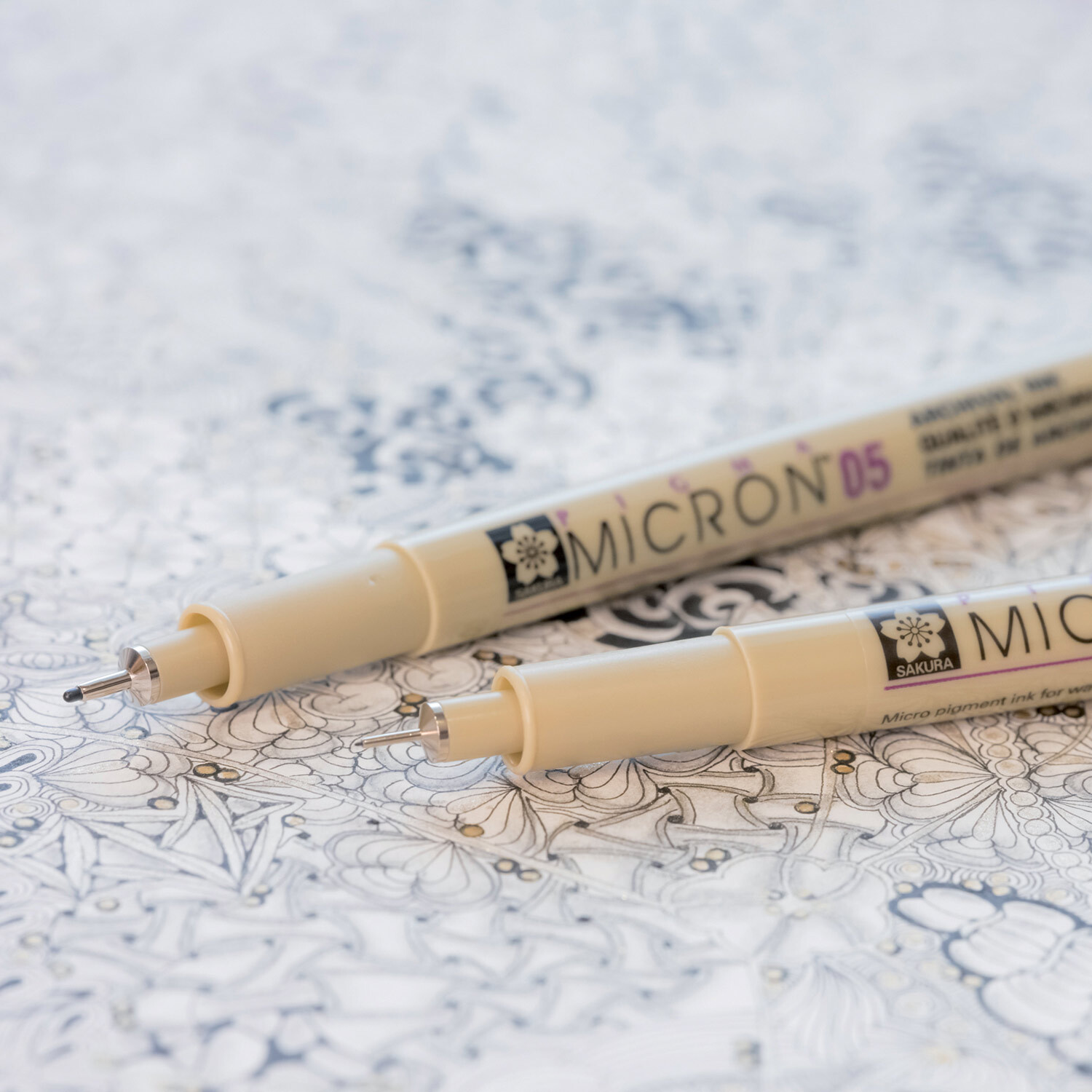 Zwei schwarze Sakura Pigma Micron Fineliner liegen auf einem detaillierten, gezeichneten Hintergrund. Die Stifte sind Teil eines 10er-Sets und eignen sich ideal für präzises Zeichnen.