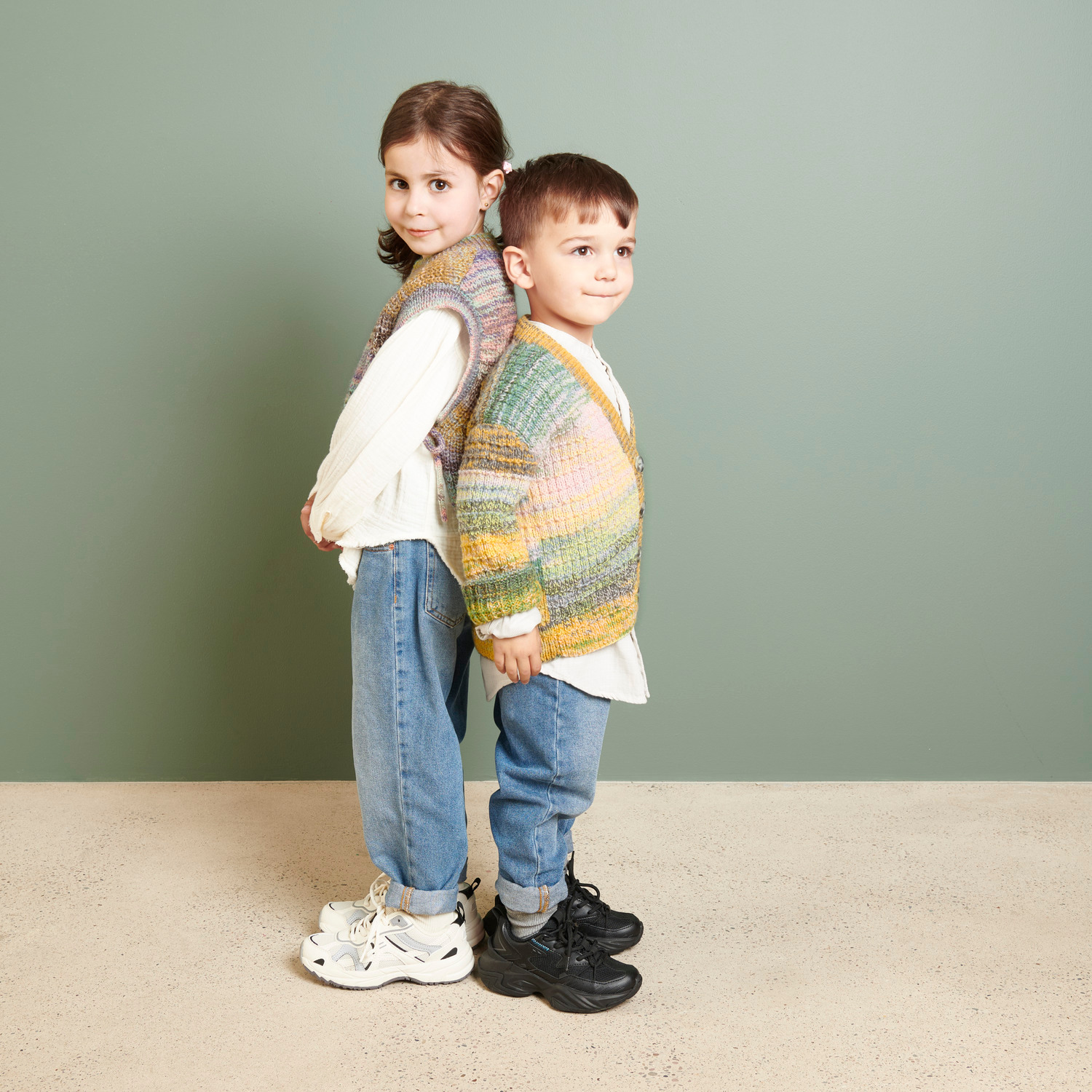 Creative Chic-Unique dk: Zwei Kinder in bunten Strickwesten vor grünem Hintergrund, lässig gekleidet.
