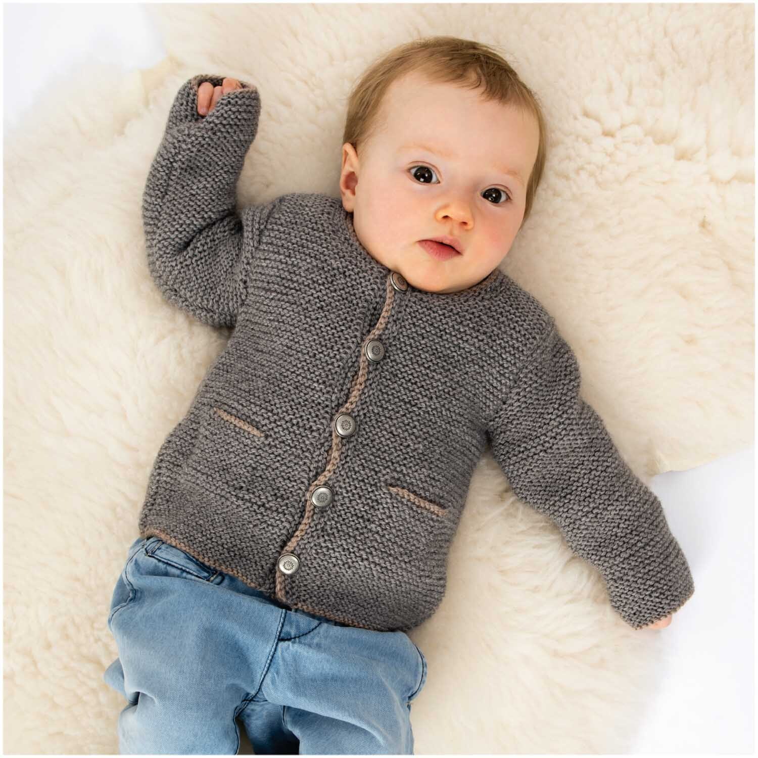 Essentials Merino Plus dk: Baby in grauer Strickjacke von Rico Design auf weicher Decke liegend.