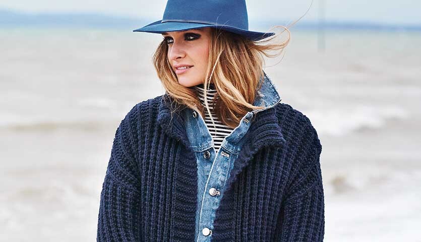 Strickanleitung Rippenmuster-Jacke aus Essentials Alpaca Blend chunky: Frau am Strand mit blauer Jacke und passendem Hut.
