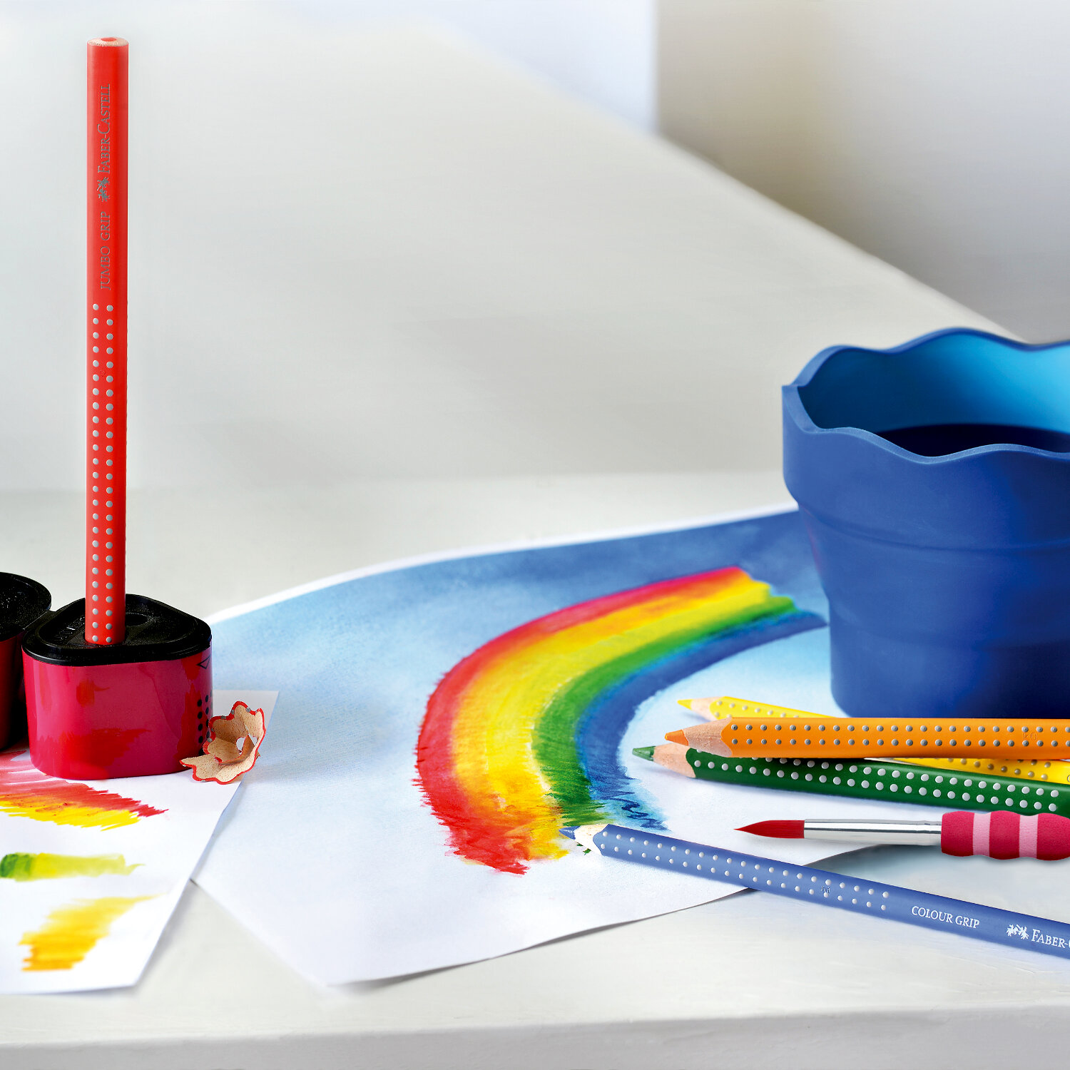 Colour Grip 2001: Bunte Stifte von Faber Castell auf einem Tisch, neben einem gemalten Regenbogen.