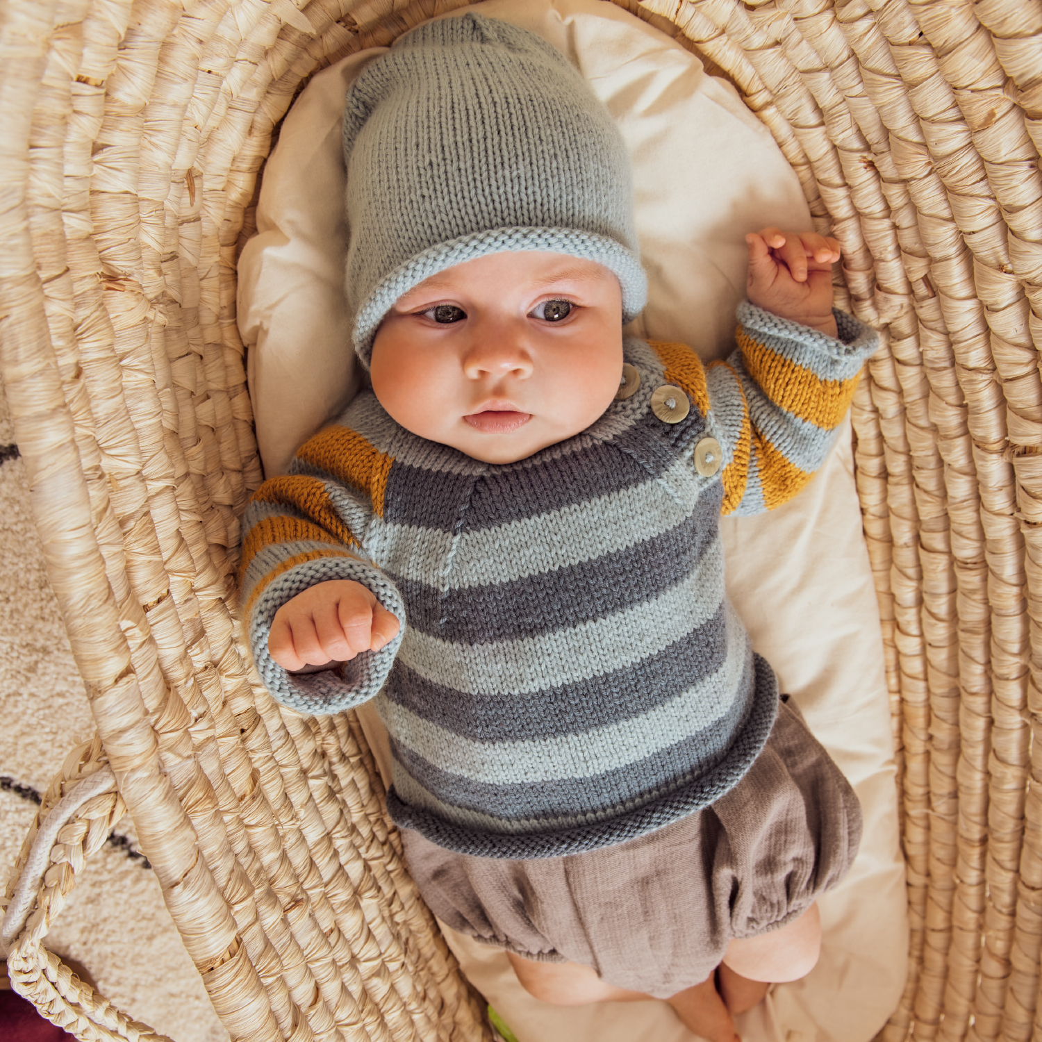 Strickanleitung Babypullover und Mütze: Baby in gestreiftem Pullover und Mütze auf geflochtener Unterlage.
