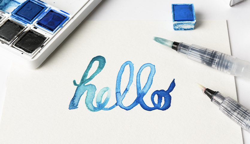 Anleitung Watercoloring Schriftzug mit der Blending-Technik: Blaues "hello" auf Papier, umgeben von Aquarellfarben und Pinseln.
