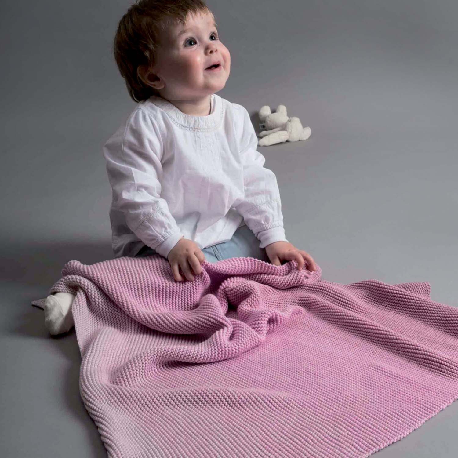 Strickanleitung Babydecke aus Creative Cotton dégradé: Ein Baby sitzt auf dem Boden mit einer rosa gestrickten Decke.