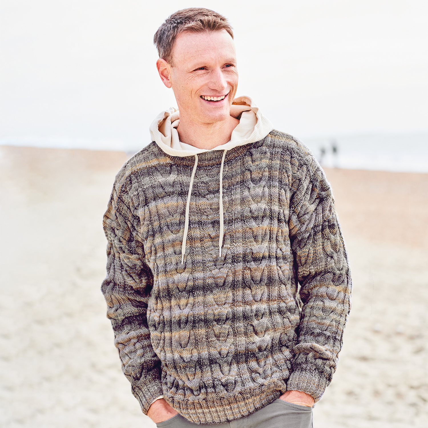 Strickanleitung Pullover aus Creative Melange aran Wonderball: Mann am Strand im gestrickten Pullover, lächelnd und entspannt.