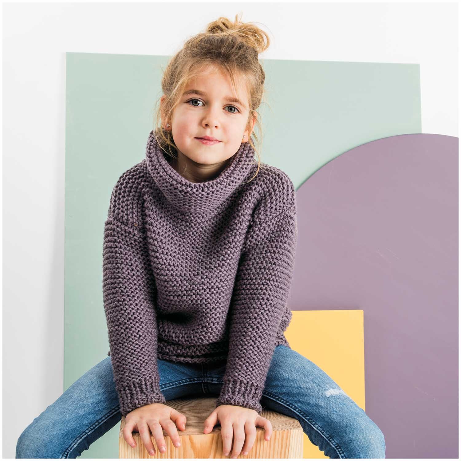 Essentials Soft Merino aran: Kind in gestricktem Pullover, sitzt auf Holzblock vor pastellfarbenem Hintergrund.