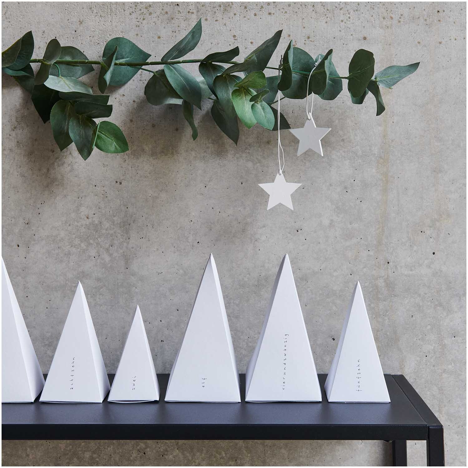 Paper Poetry Adventskalender Bäume 24 Stück, weiße Pyramiden auf schwarzem Regal vor Betonwand.