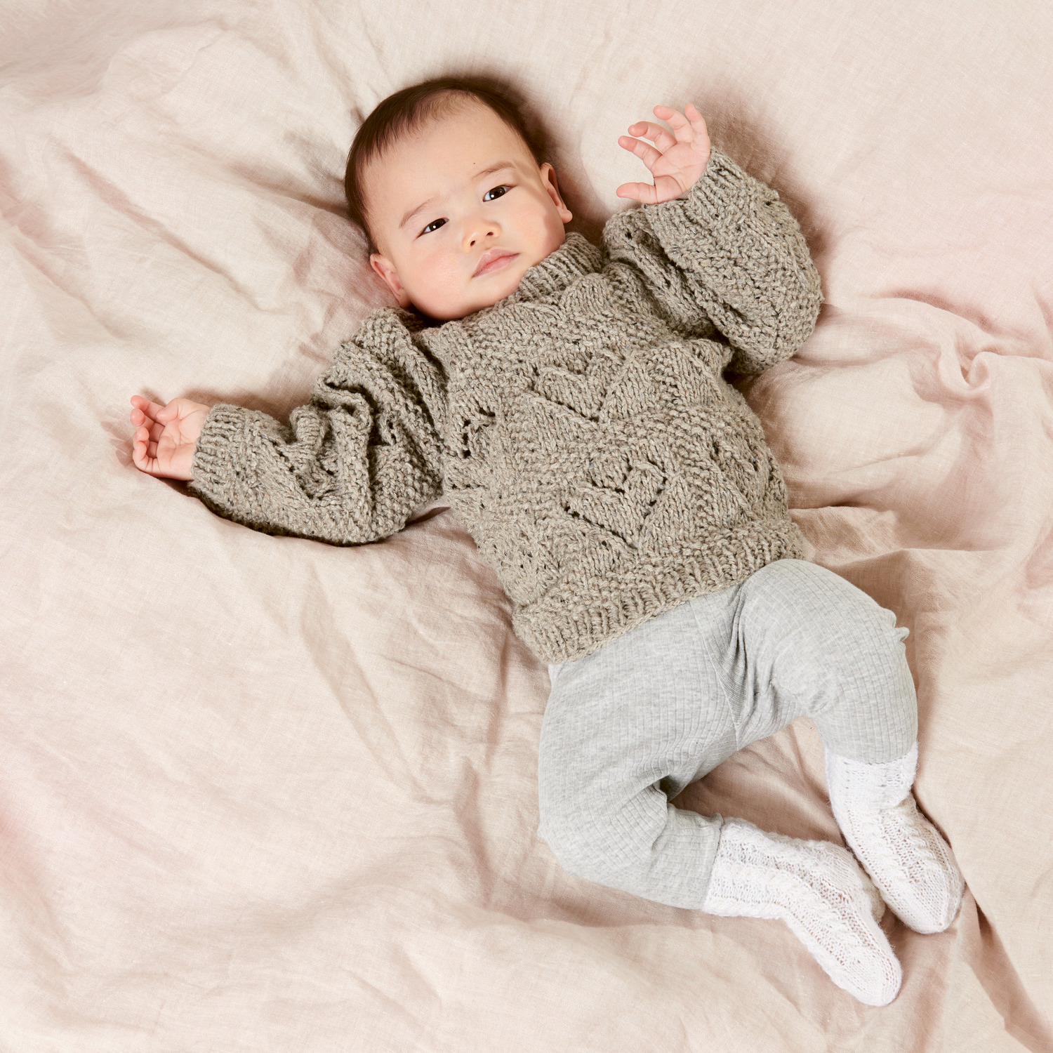 Strickset Babypullover Modell 06: Baby in grauem Pullover mit Herzmuster, liegend auf beiger Decke.
