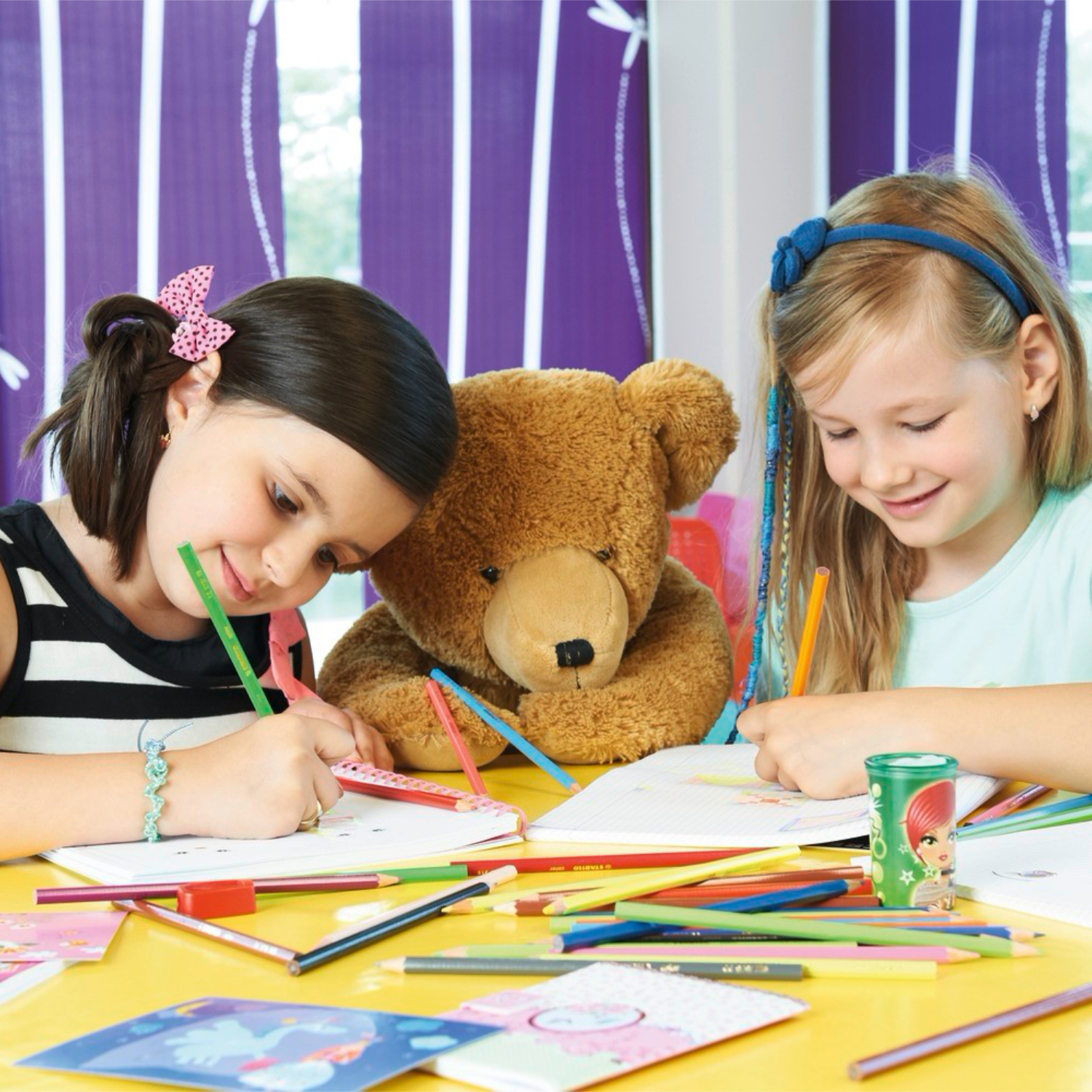 STABILO color Buntstifte: Zwei Kinder malen fröhlich mit Buntstiften an einem Tisch, begleitet von einem Teddybär.